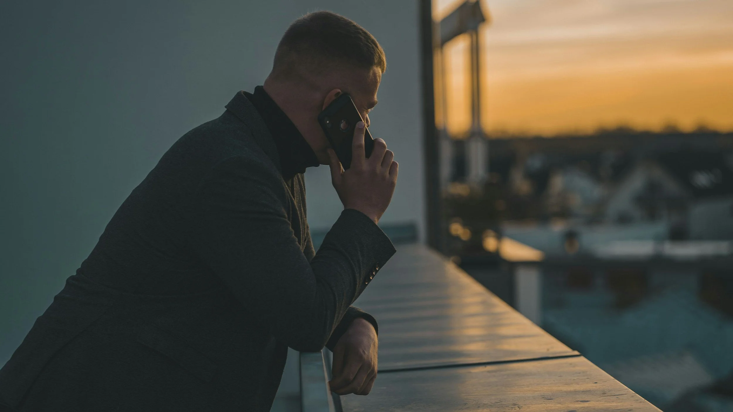 A man in a dark jacket is talking on his cellphone, leaning on a railing with a cityscape and sunset in the background.