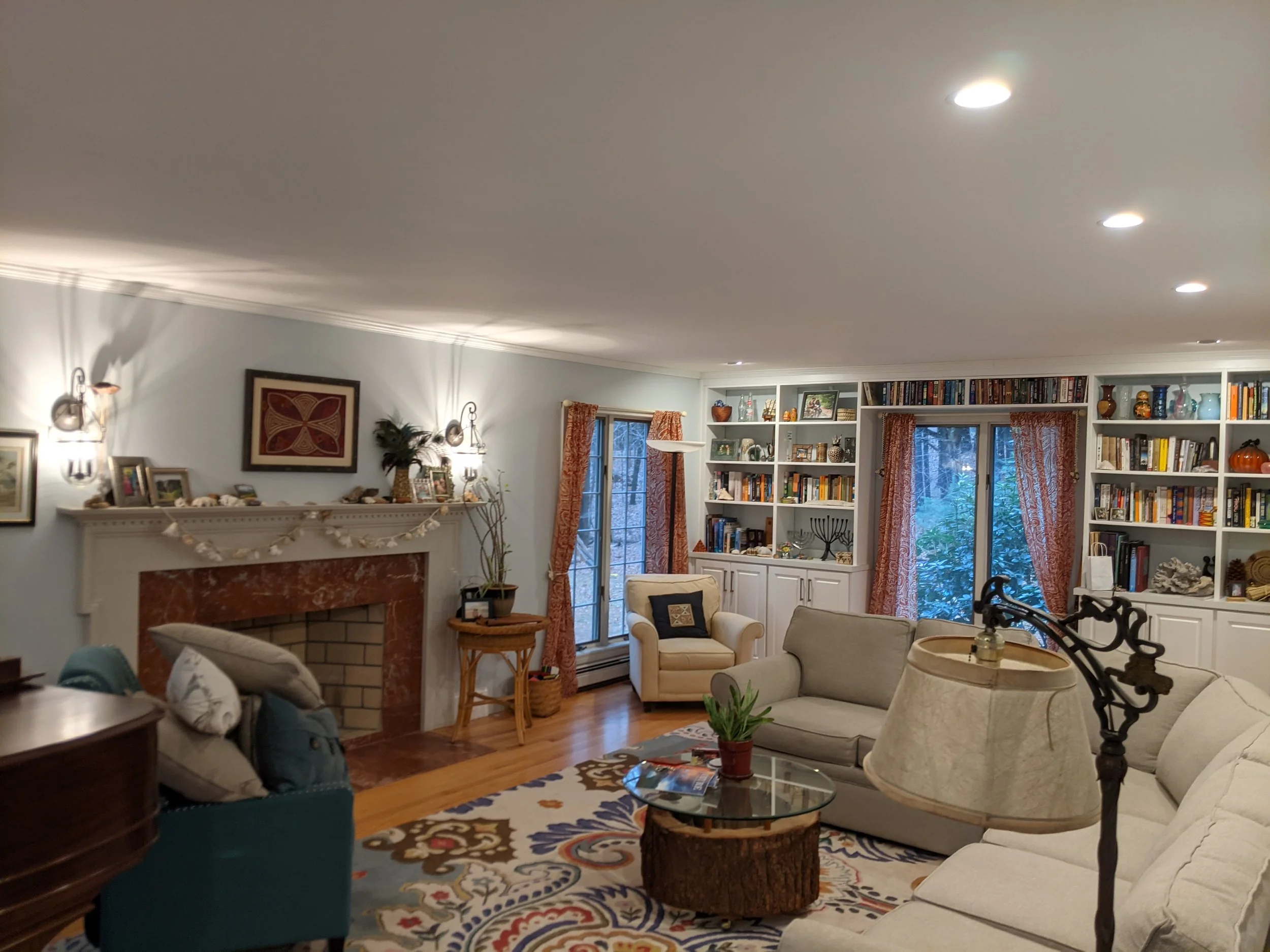 Living room with white couch and armchair, fireplace with red marble surround, built-in bookshelves, windows with orange curtains, floor lamp, and decorative items.