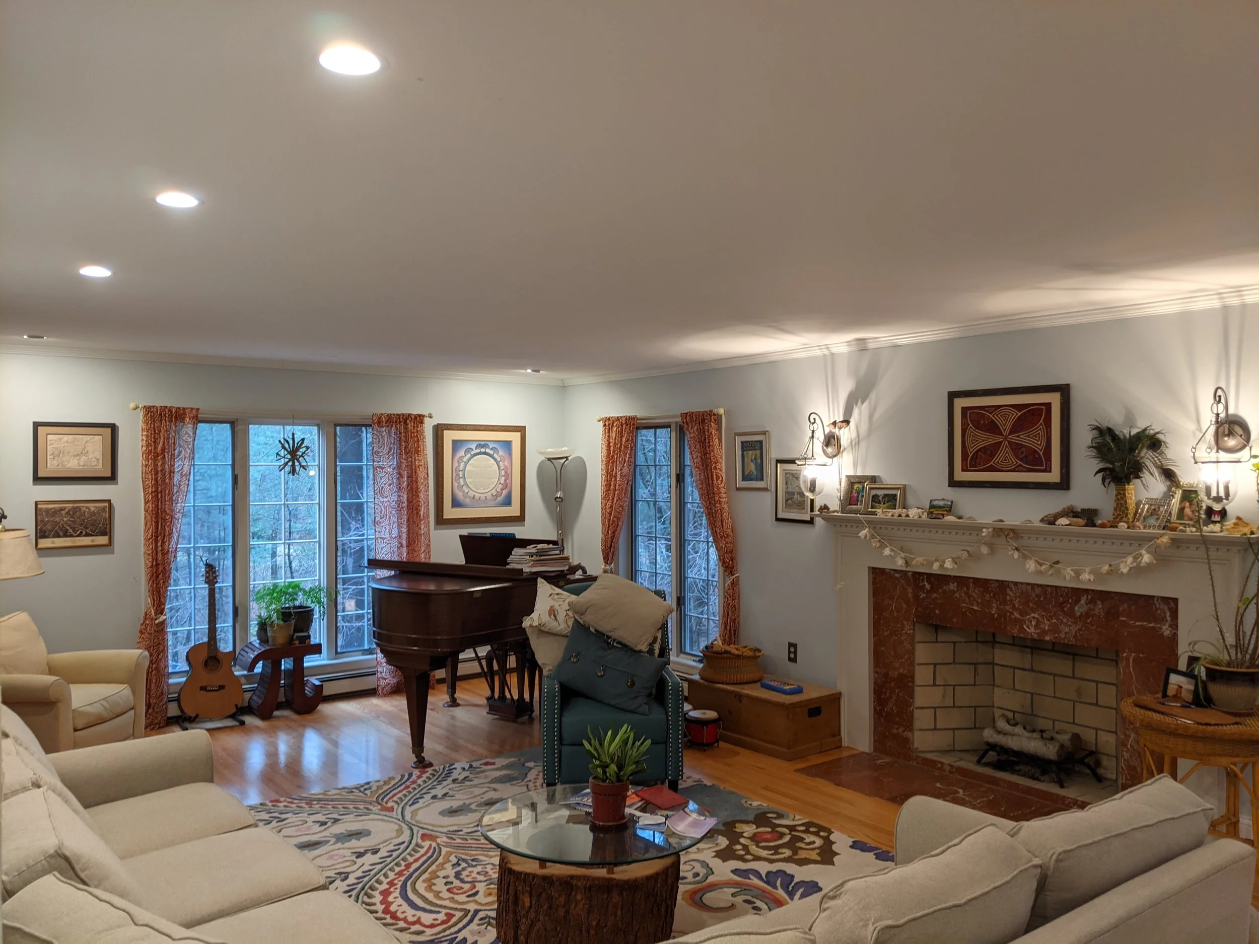 A cozy living room with a beige sofa, a glass-topped coffee table, and an armchair with pillows. There is a fireplace with a red marble surround and various decorations on the mantel. The room has large windows with orange curtains, a grand piano, an