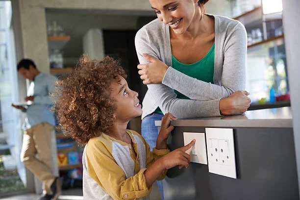 A woman and a young girl are smiling and talking in a kitchen. The girl is pointing at an electrical outlet on the counter, and the woman is leaning on the counter, looking at her.