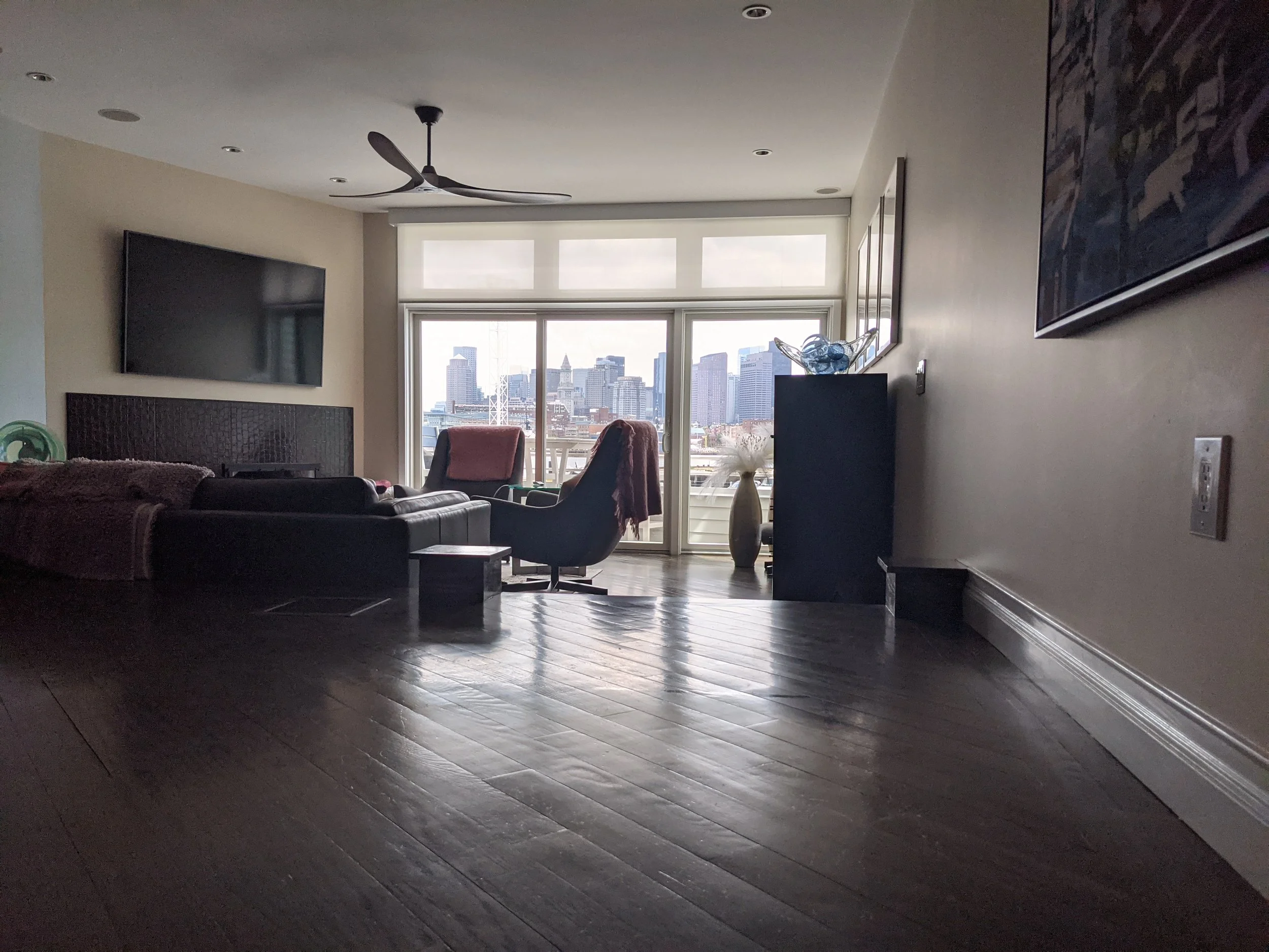 Living room with dark wooden floor, black leather couches, armchairs with blankets, flat-screen TV on the wall, large windows with cityscape view, ceiling fan, decorative vases, and abstract wall art.