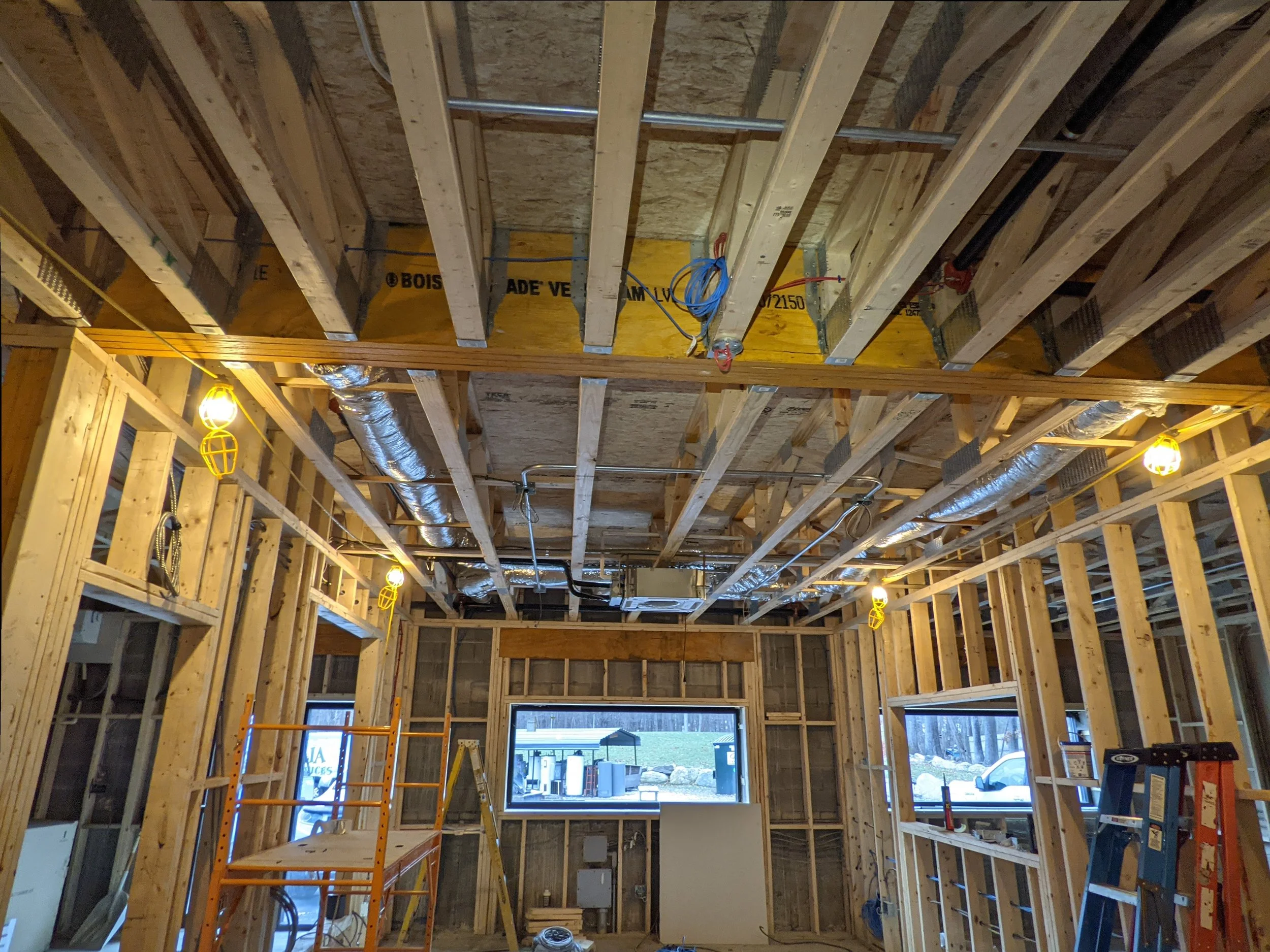 Interior of a house under construction with exposed wooden framing, electrical wiring, and HVAC ductwork. Temporary lighting fixtures illuminate the workspace.