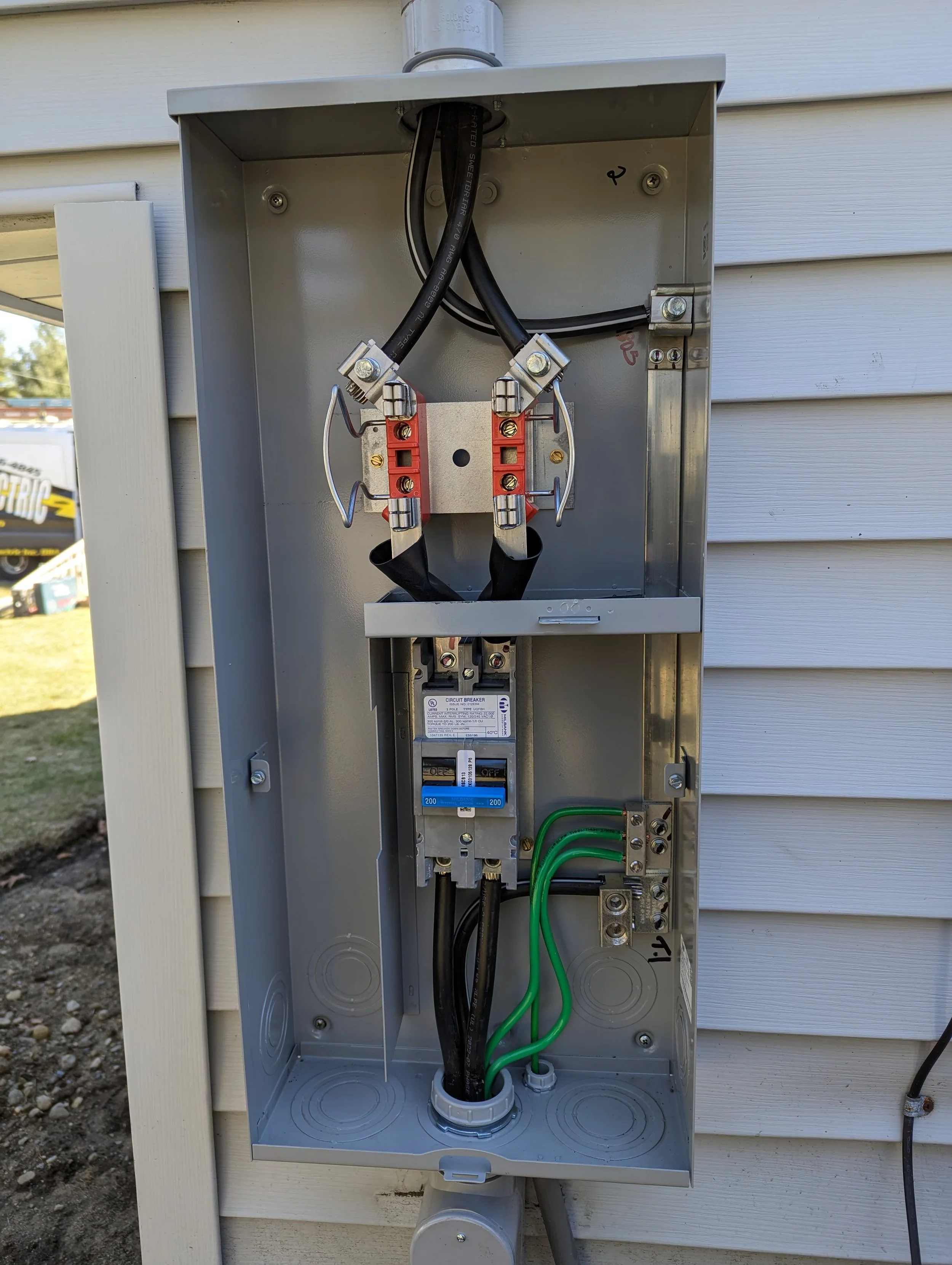 Open electrical service panel with black and green wires, a circuit breaker, and metal components inside a gray box mounted outside on a house wall.