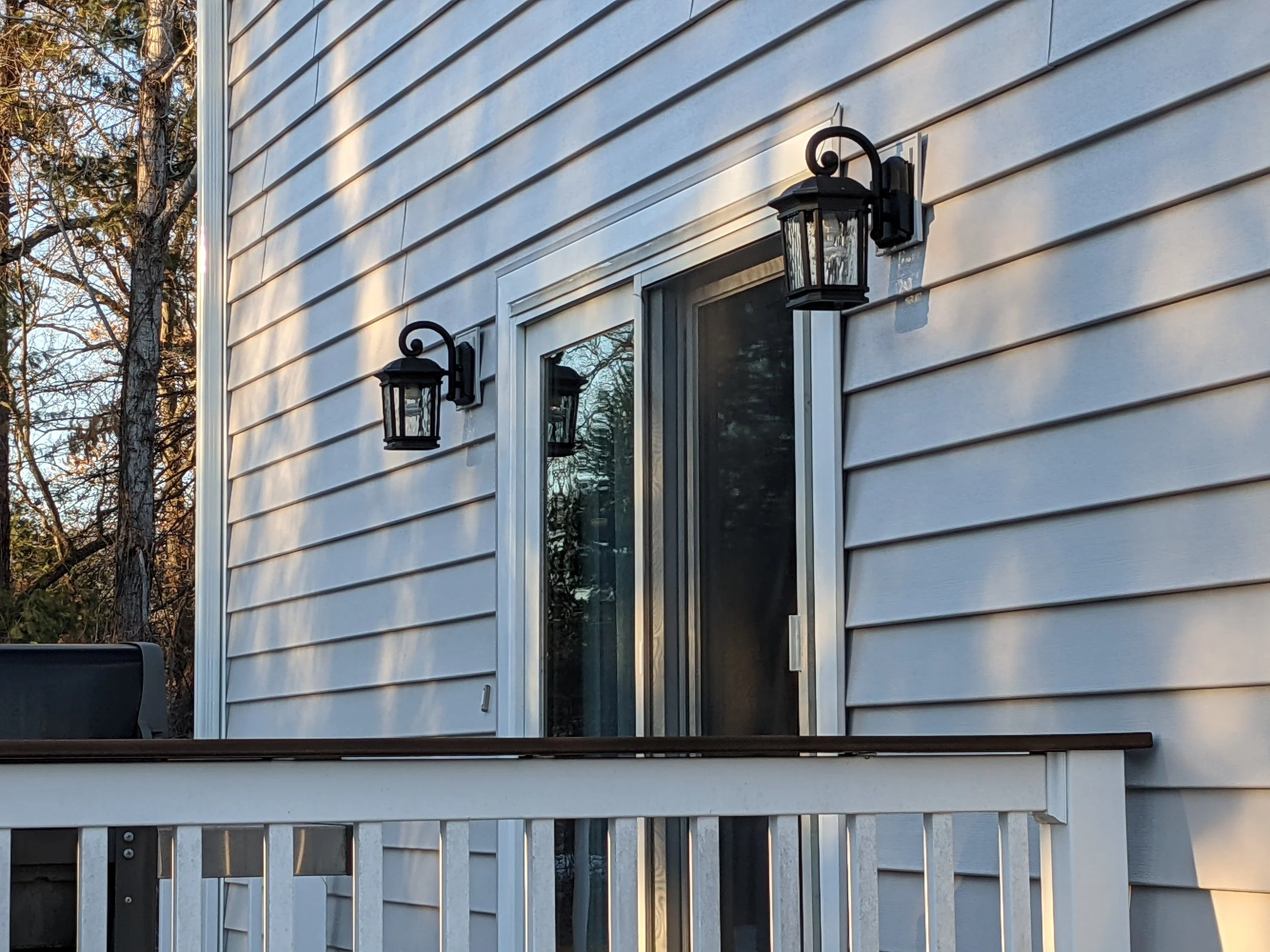 Exterior of a house with white vinyl siding, two black outdoor lantern lights, a sliding glass door, and a white deck railing, with trees in the background.