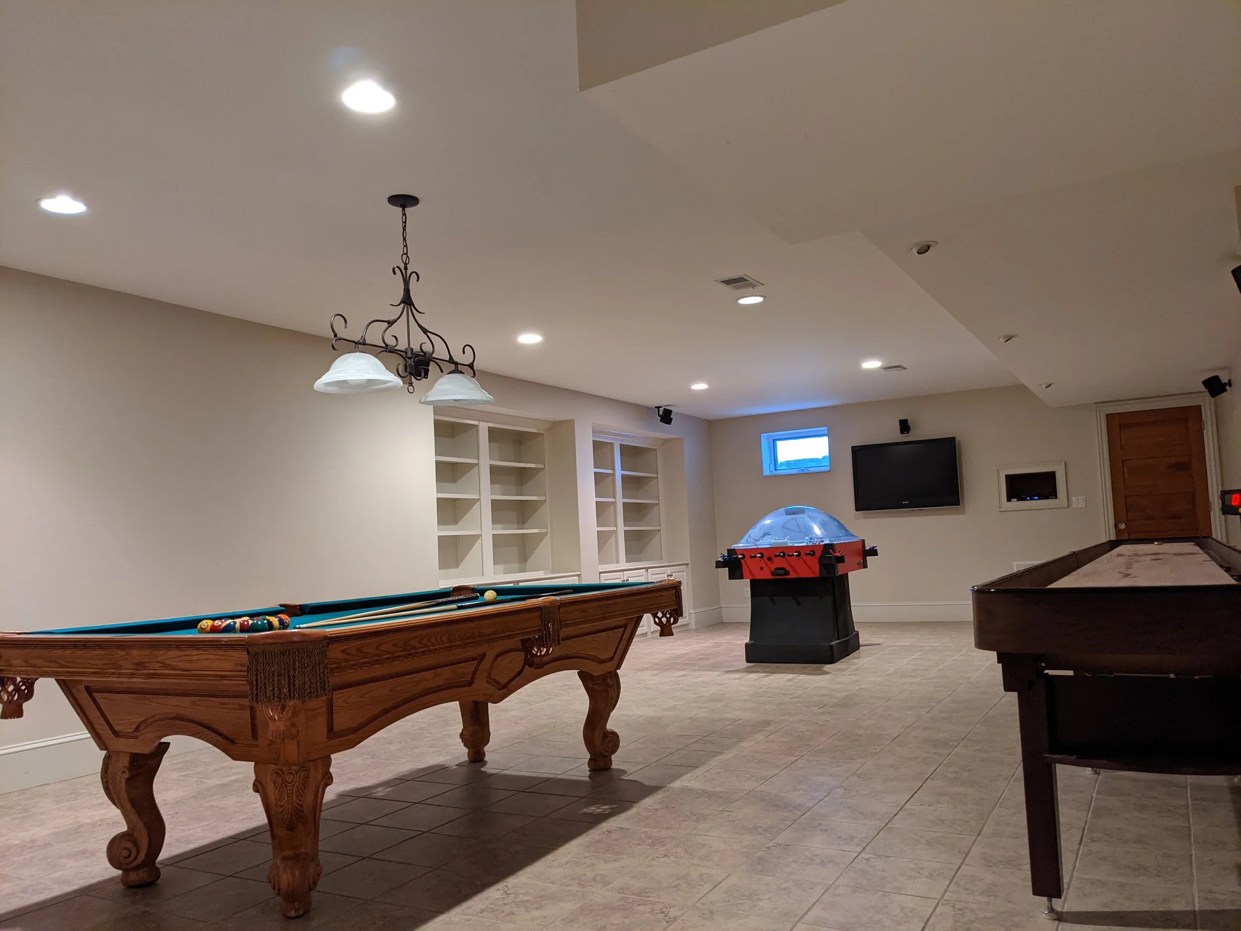 Game room with pool table, air hockey table, and foosball table, with built-in shelves, television, and small window on beige walls and tiled floor.