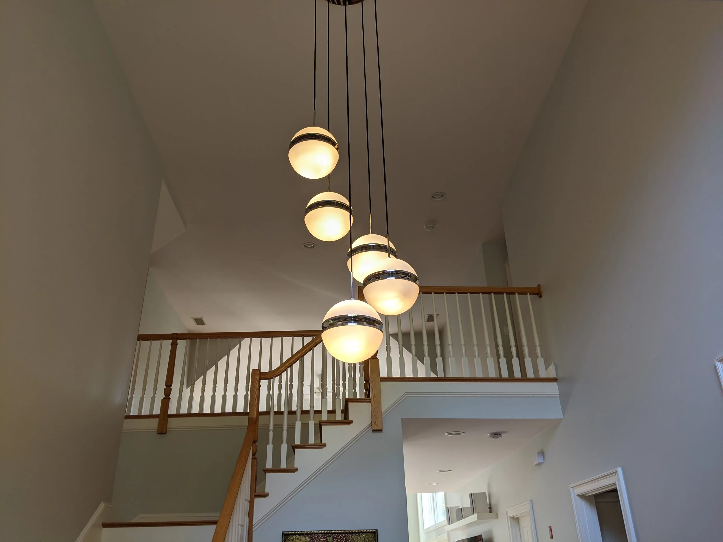 A modern chandelier with six spherical light fixtures hanging from a high ceiling in a house interior with a staircase and railing.