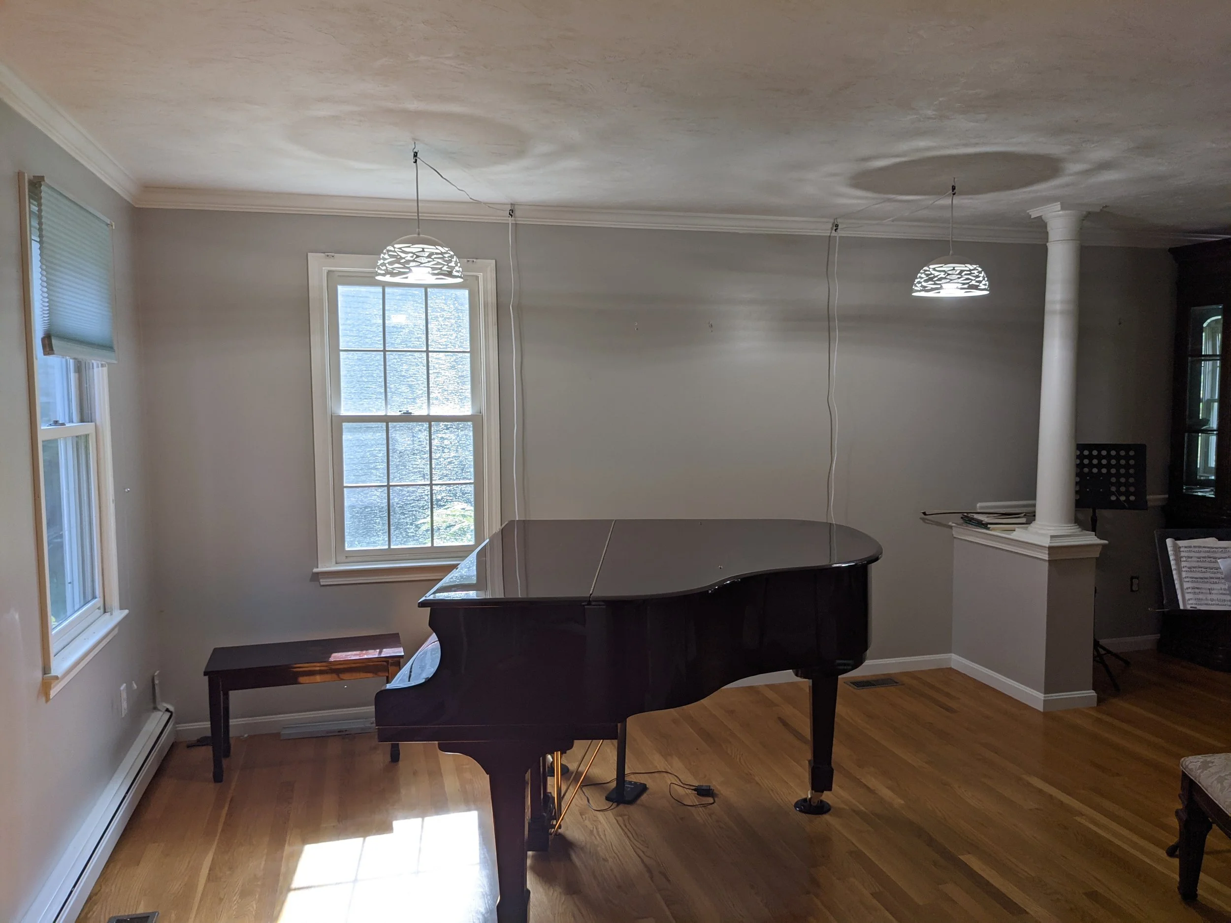A black grand piano in a bright living room with hardwood floors, two windows with blinds, and contemporary hanging lights.