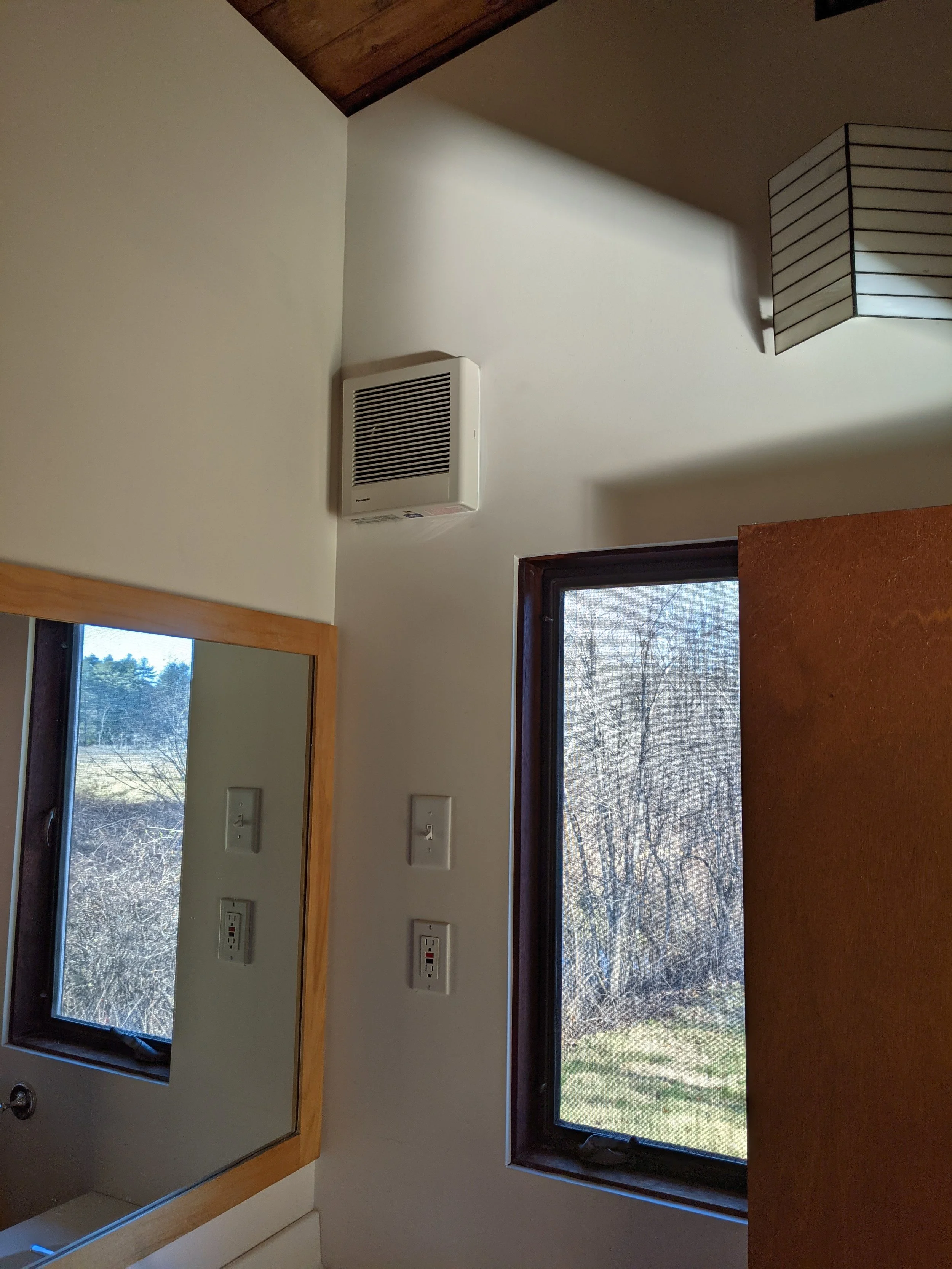 Interior view of a room showing a window, a wall-mounted white fan or vent, multiple electrical outlets, and a mirror reflecting outdoor scenery with trees.
