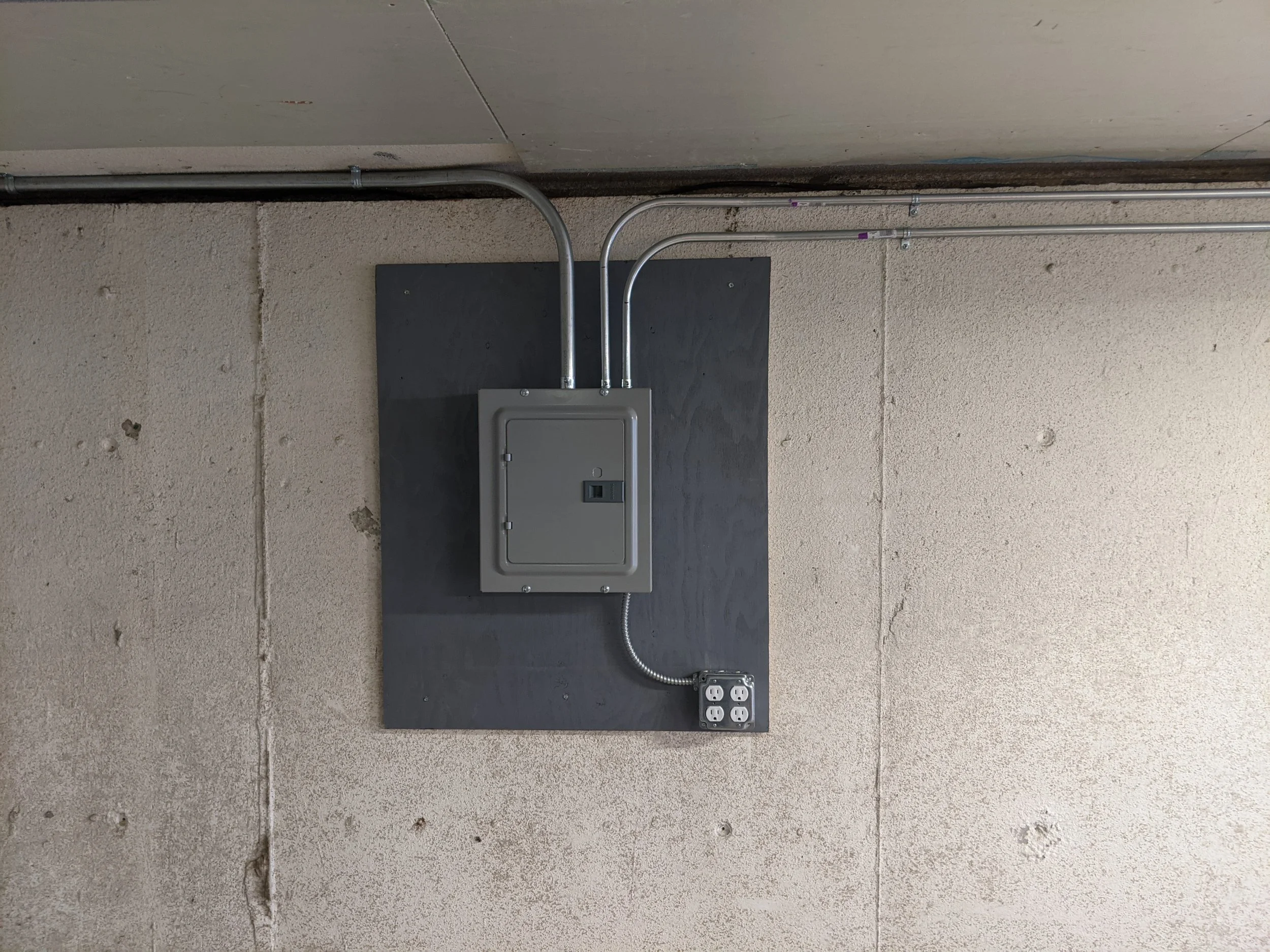 Electrical meter box on a black mounting board on a beige concrete wall with wiring and conduits.