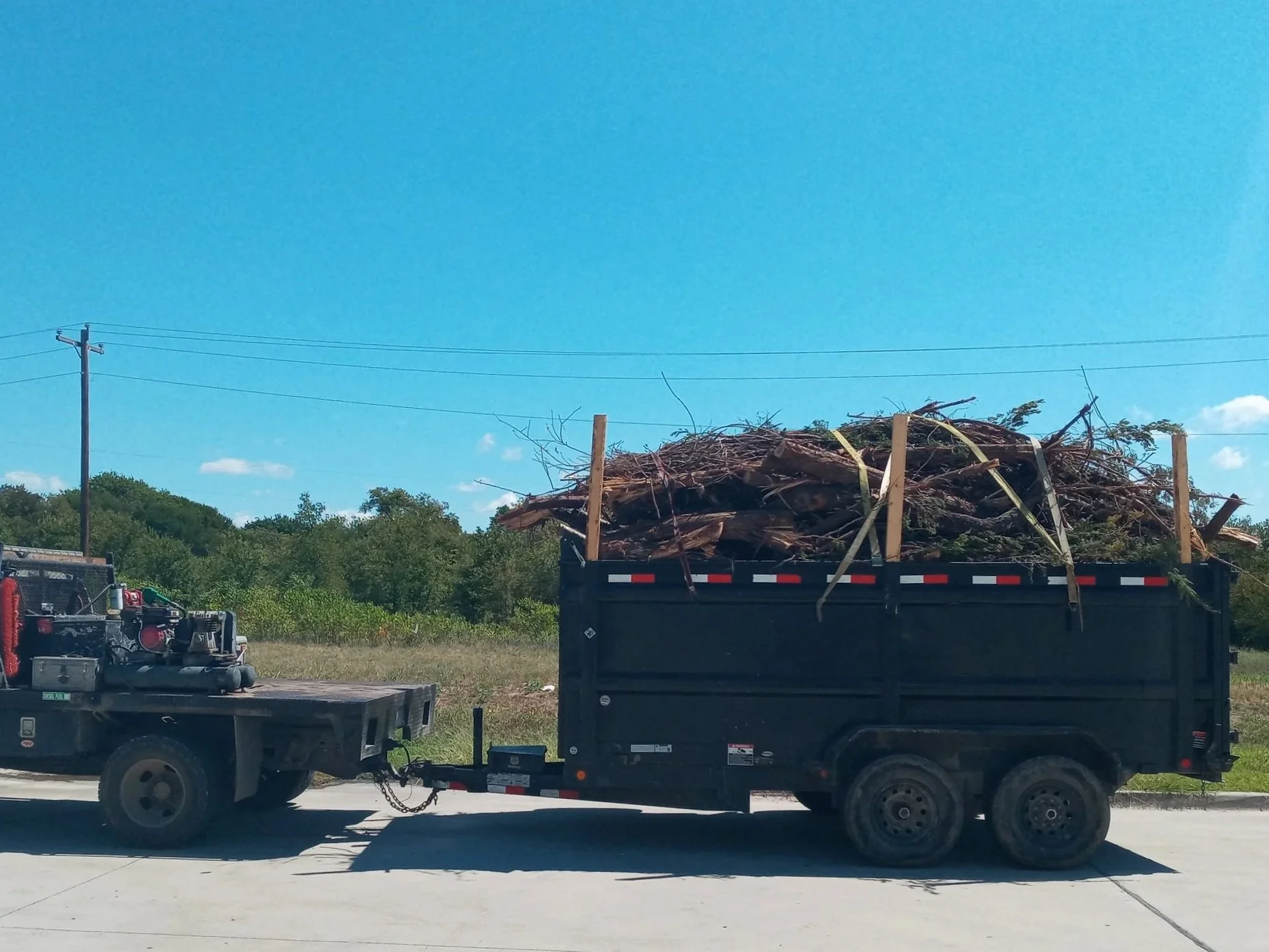 Black trailer filled with branches and logs, attached to a flatbed truck on a sunny day with a blue sky and green trees in the background.