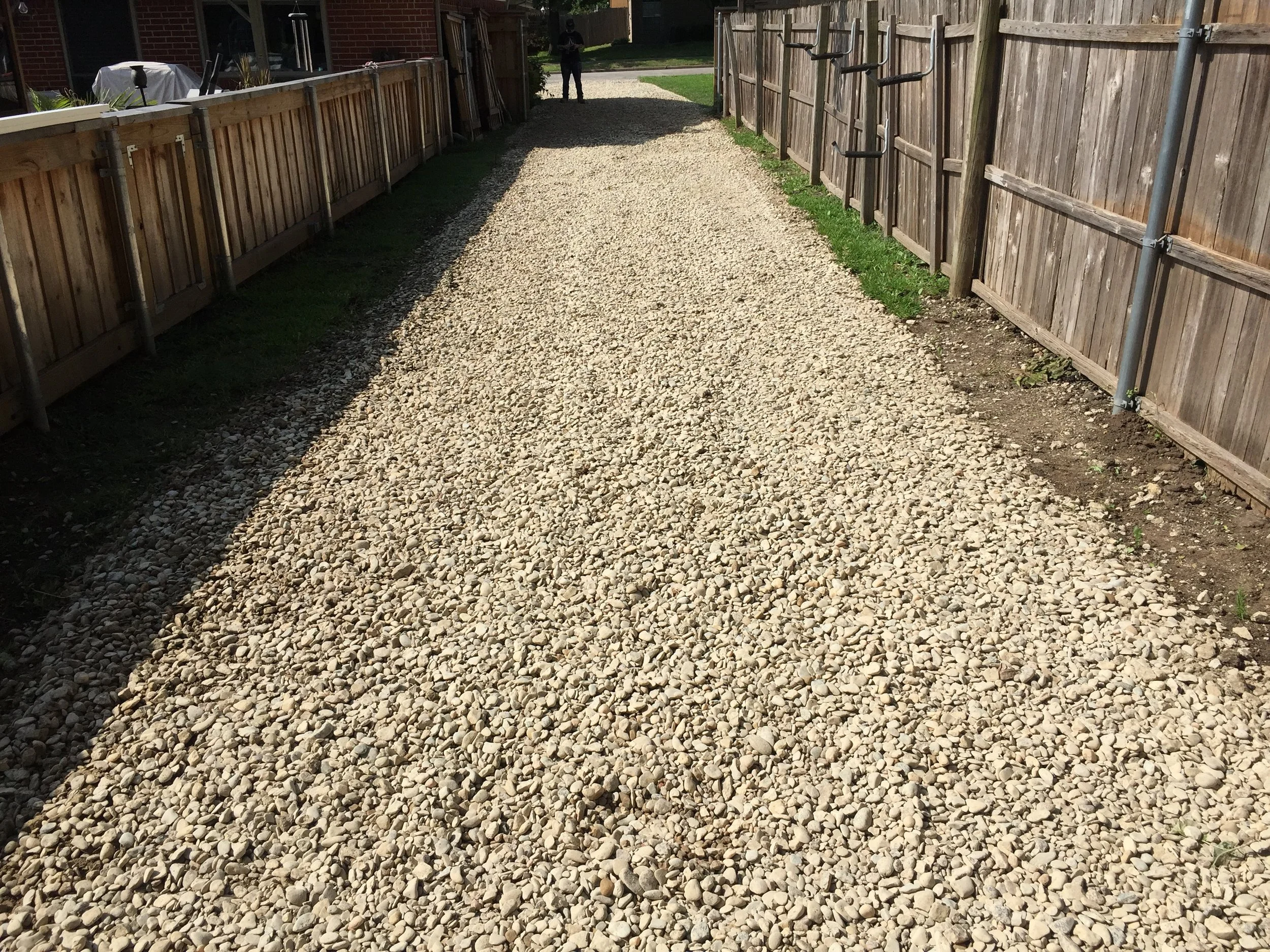 A gravel pathway between two wooden fences, with a person walking in the distance.