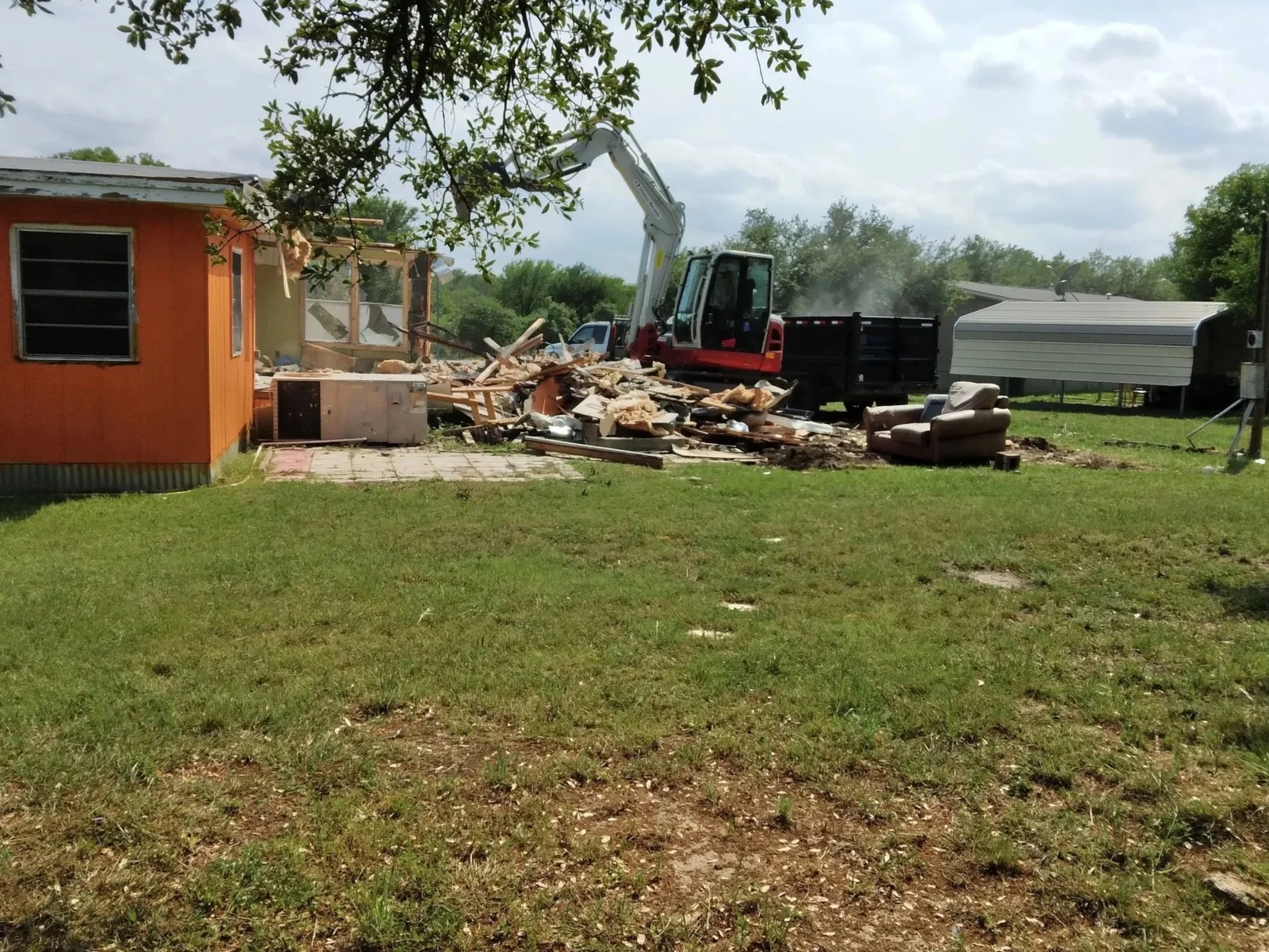 A house is being demolished with a small excavator, debris scattered around, a green lawn, a brown armchair outside, and a mobile home in the background under a cloudy sky.