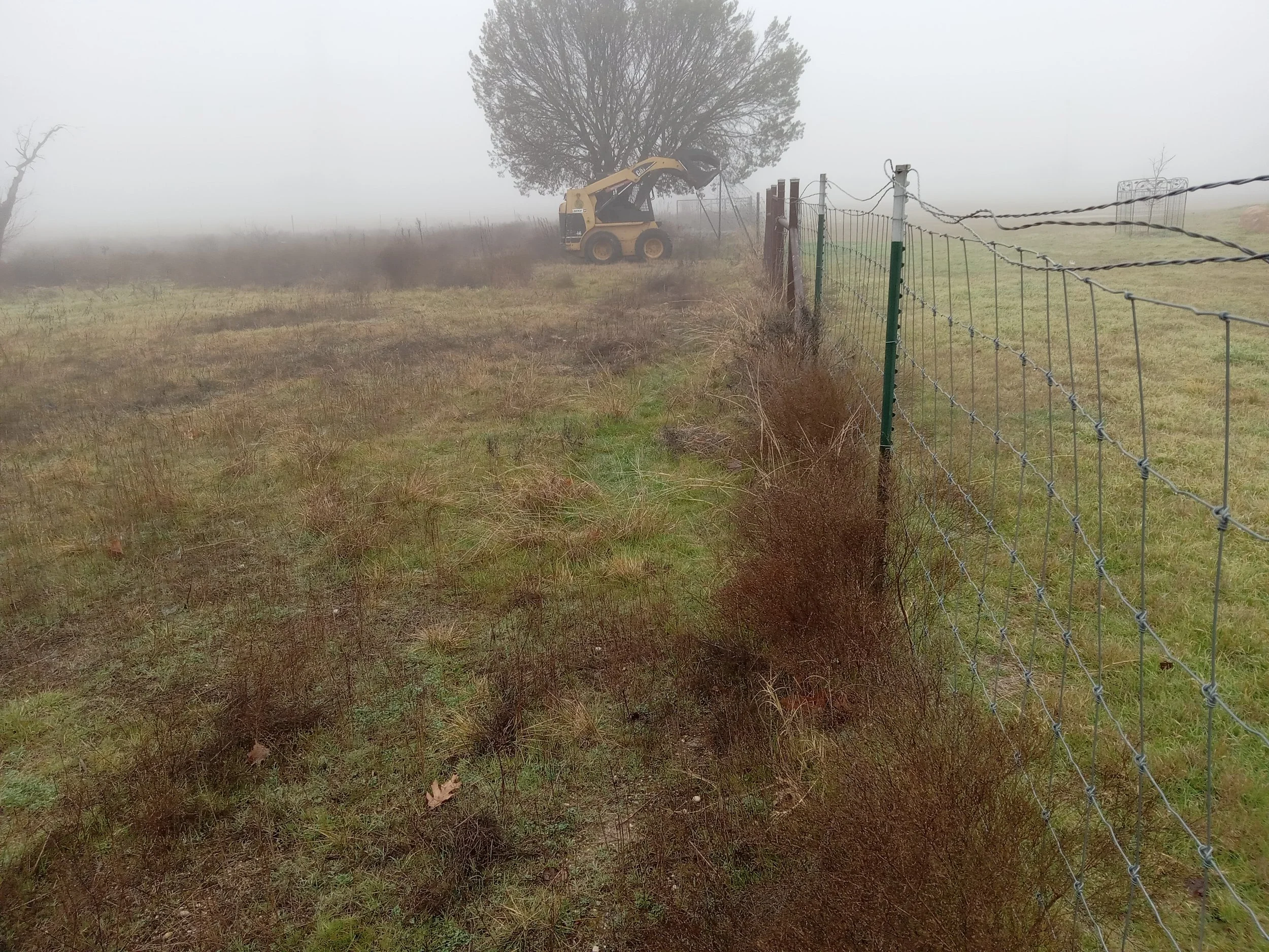 A foggy rural landscape with a barbed wire fence on the right side, a tree in the background, and a small bulldozer working near the fence in the distance.