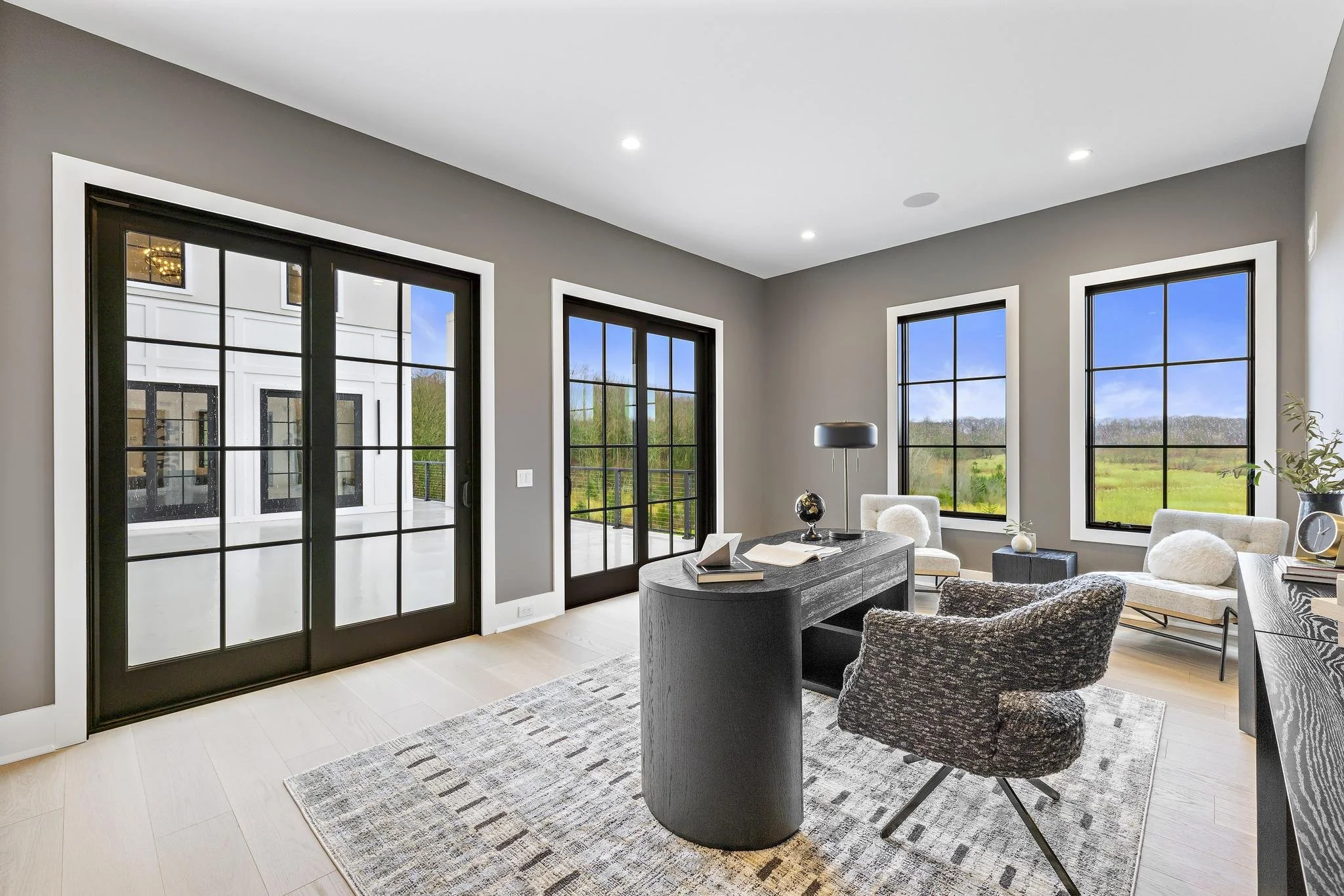 Modern home office with large windows showing outdoor landscape, gray walls, black framed glass doors, light wood flooring, and minimalist furniture including a black desk, patterned office chair, and white seating.