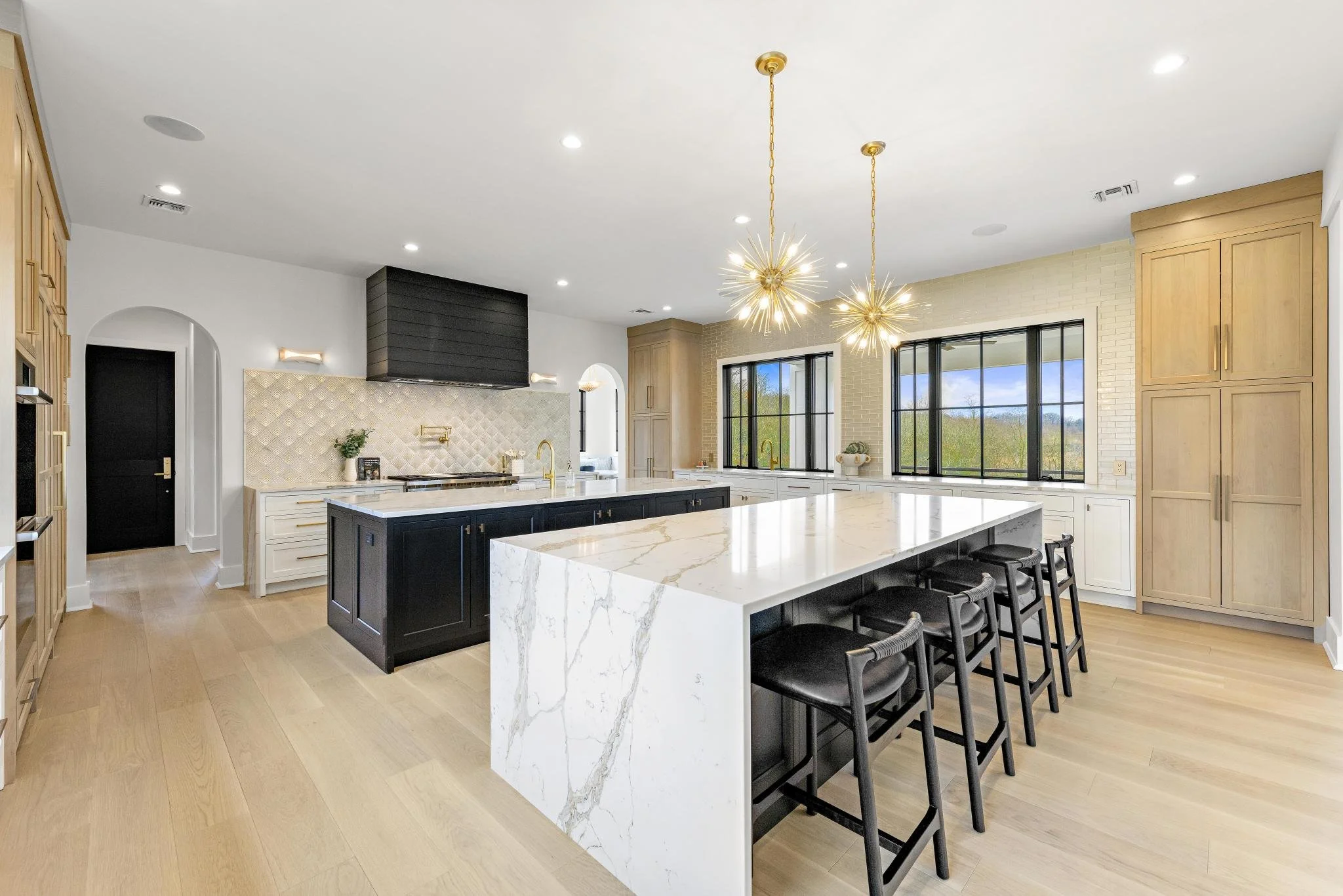 Modern kitchen with black and white cabinetry, marble island, large windows, and gold light fixtures.