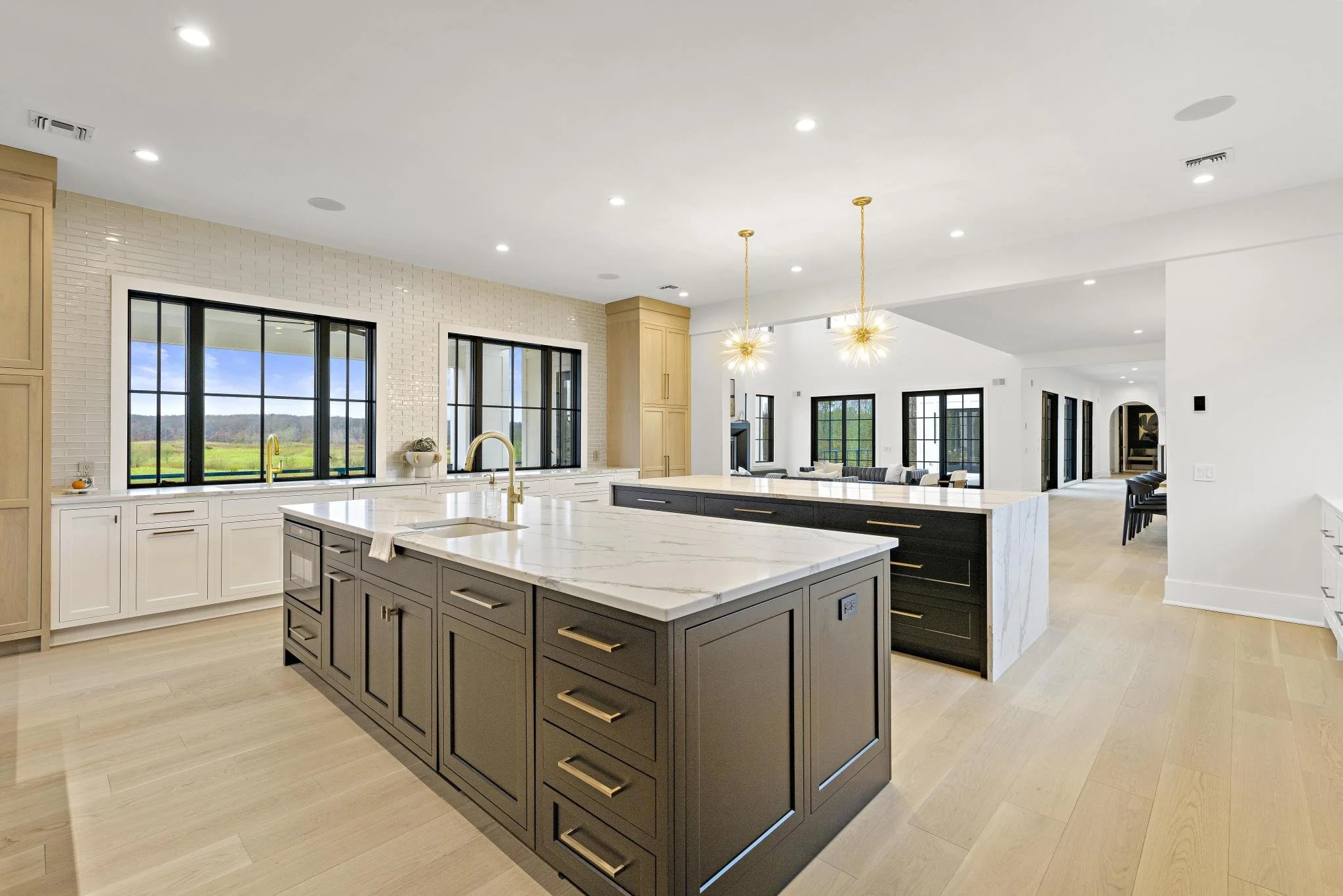 Modern open-concept kitchen with black and white cabinetry, marble countertops, large island, and pendant lighting, overlooking a spacious living area with black-framed windows and patio doors.