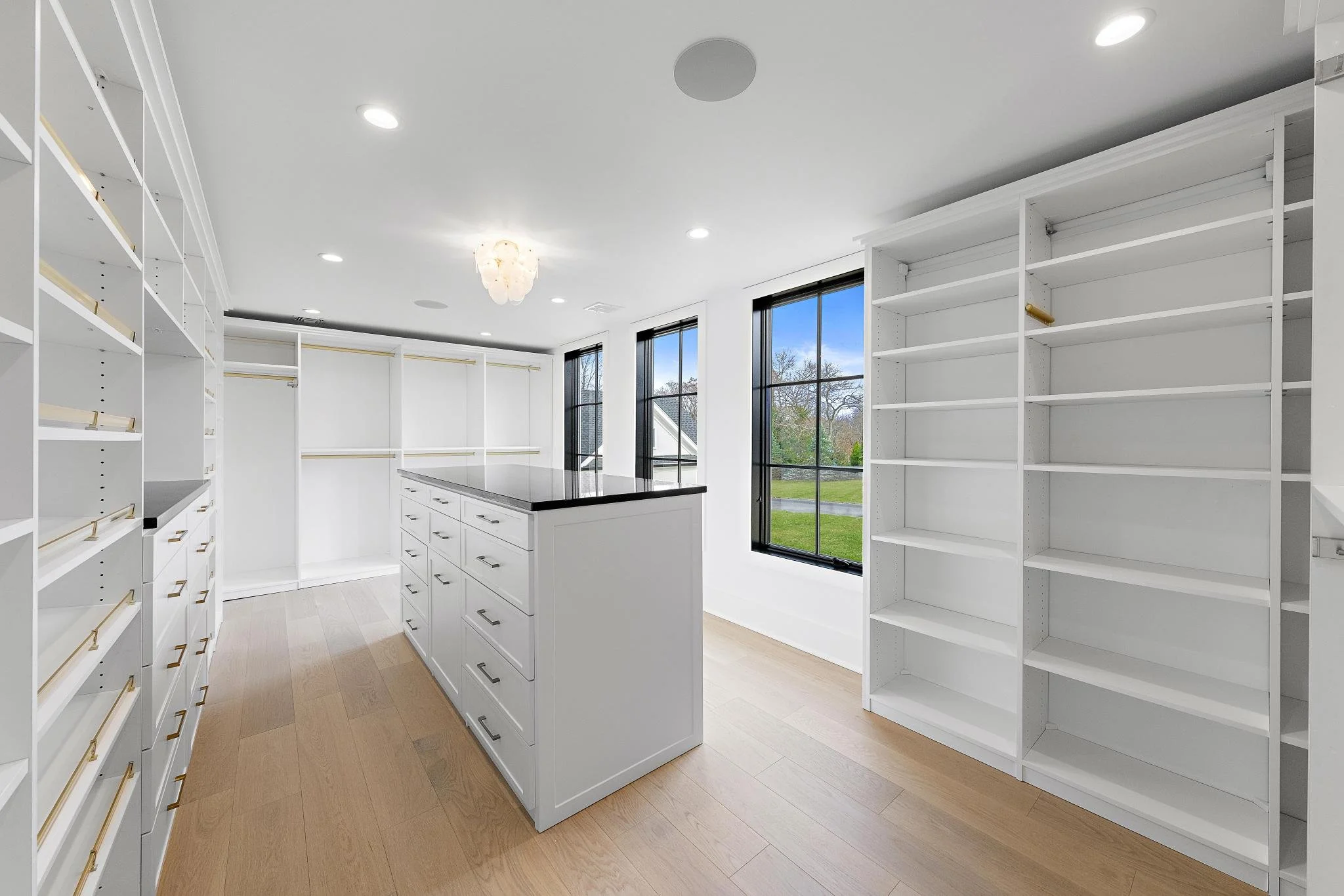 Empty walk-in closet with white built-in shelves and drawers, large windows showing outdoors, hardwood floors, and ceiling lights.