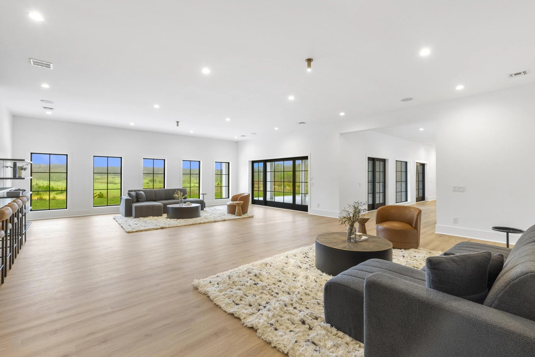Spacious living room with white walls, wooden flooring, multiple windows, and minimal furniture including sofas and chairs.