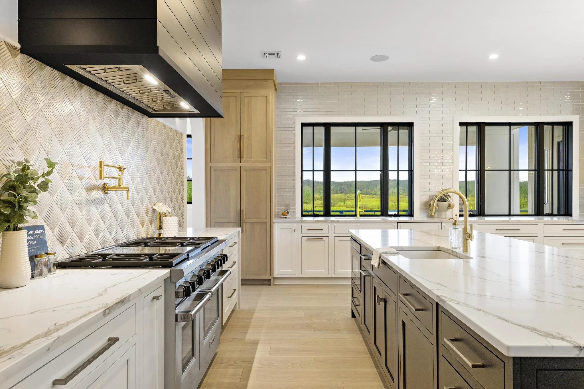Modern kitchen with white and gray cabinets, marble countertops, black window frames, and a view of green fields outside.