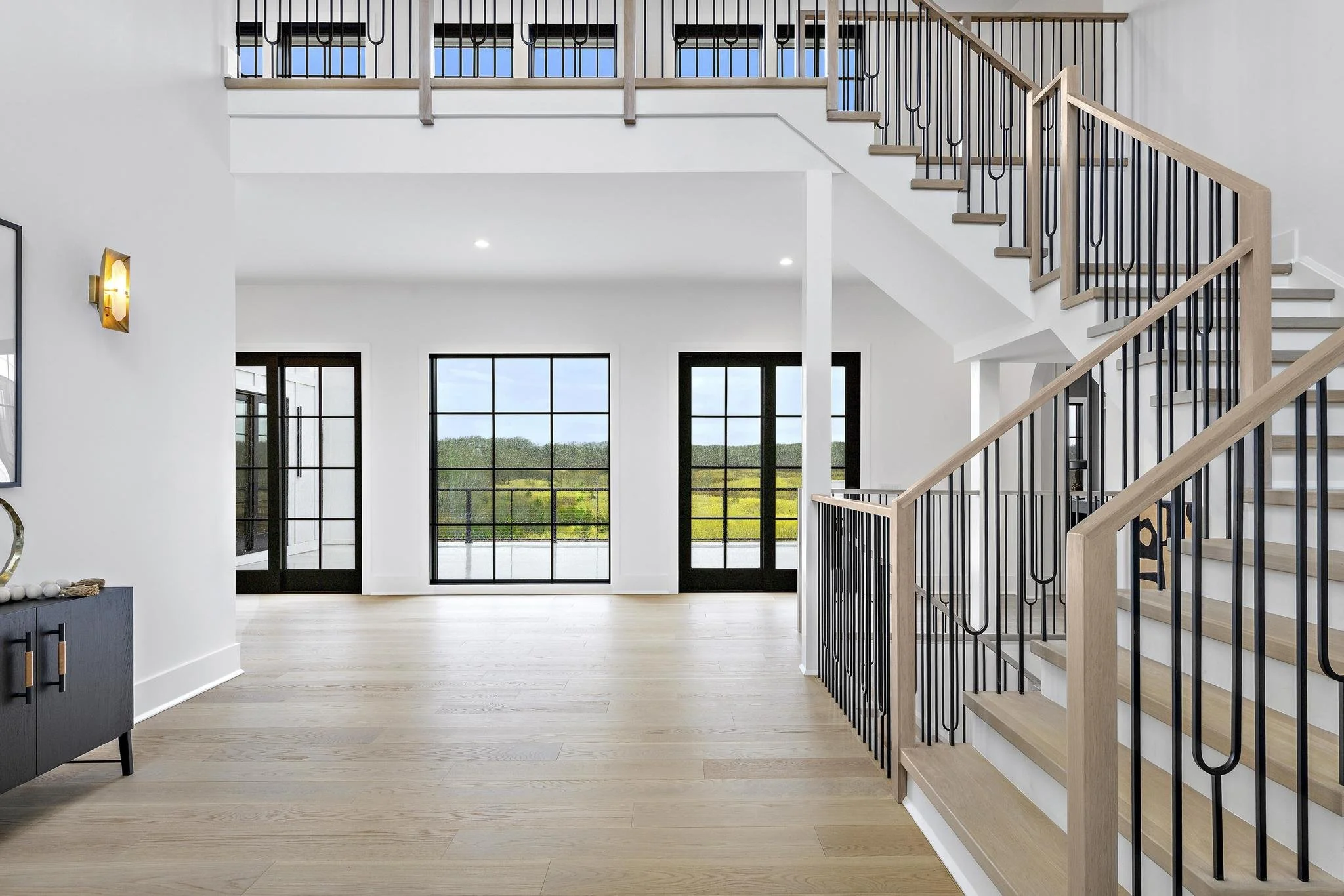 Interior view of a modern house with light wood floors, white walls, black-framed glass doors, and staircase with wooden steps and black metal railings leading to an upper level