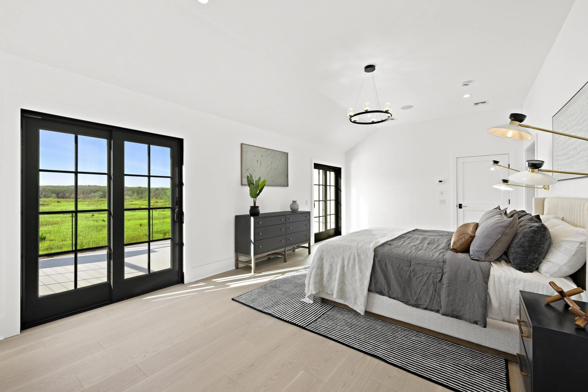 Modern bedroom with black framed glass doors leading to a patio, a bed with gray and white bedding, black nightstand, black dresser with decorative items, and large windows allowing natural light.