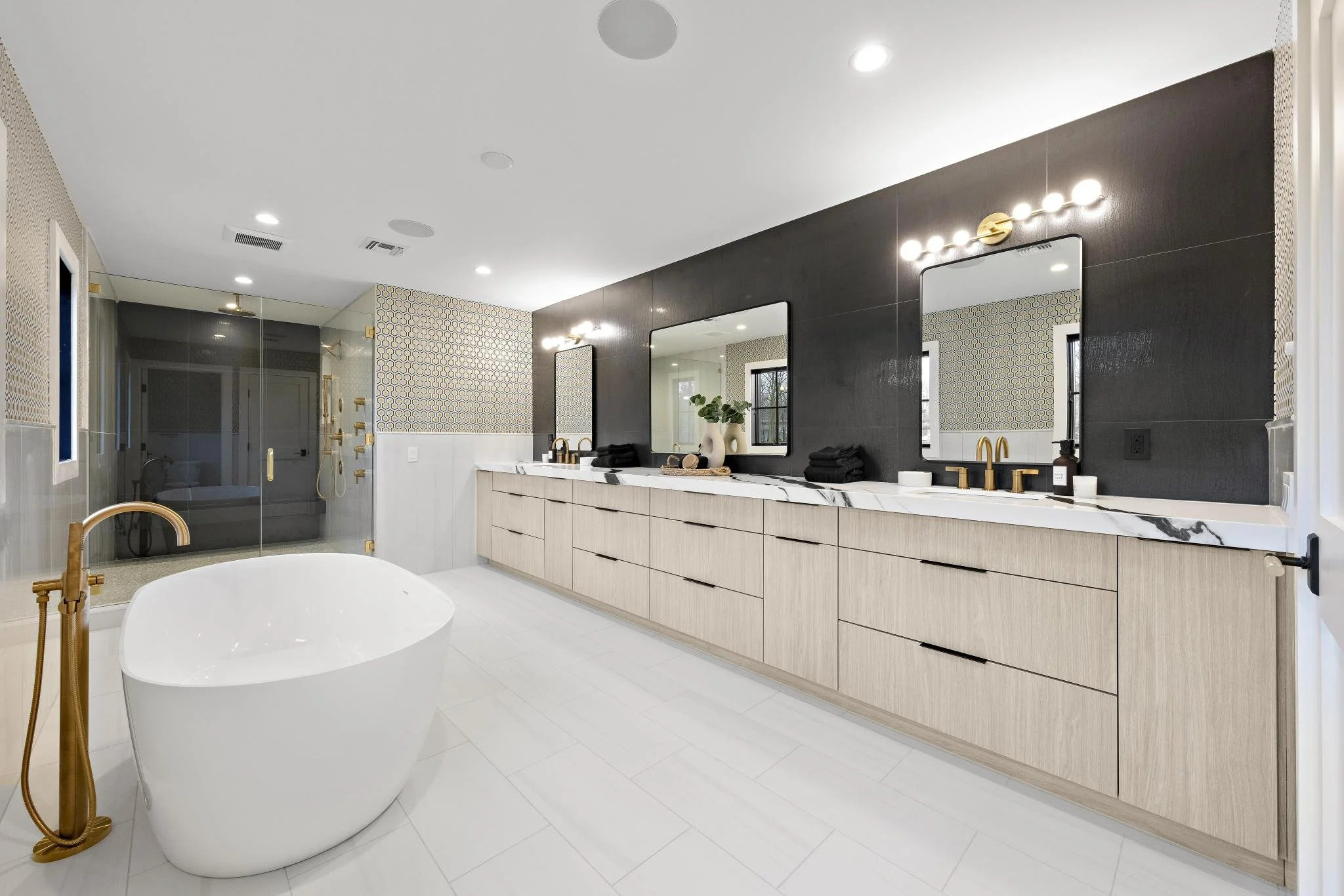 Modern bathroom with a freestanding white tub and a double vanity with gold fixtures, large mirrors, and black and white countertops. There is a walk-in shower with glass doors and a patterned accent wall.