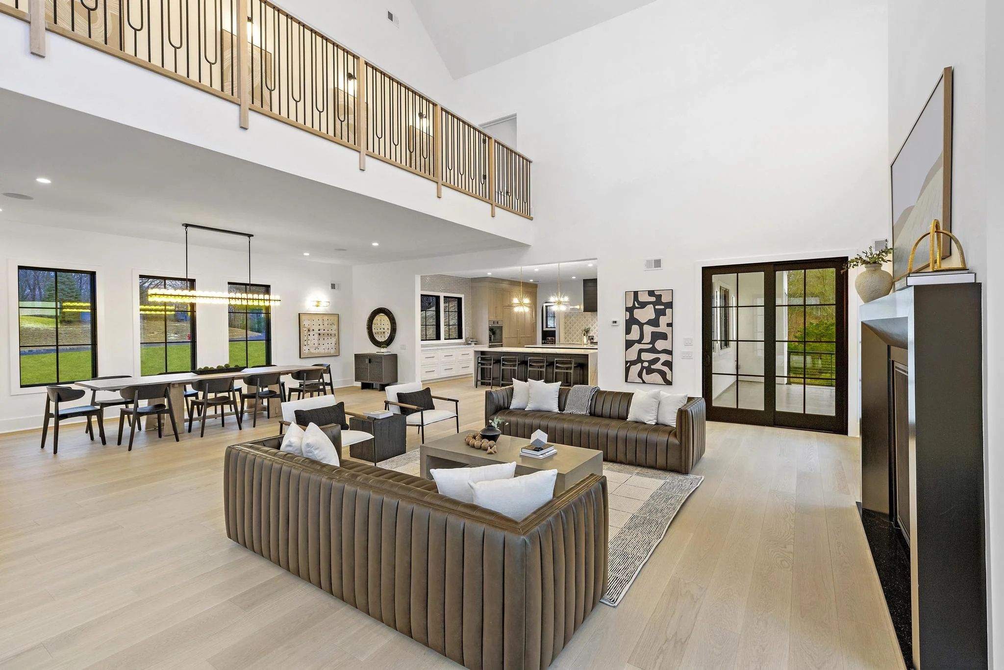Open-concept living room and dining area with high ceilings, large windows, modern furniture, and a view of the kitchen.