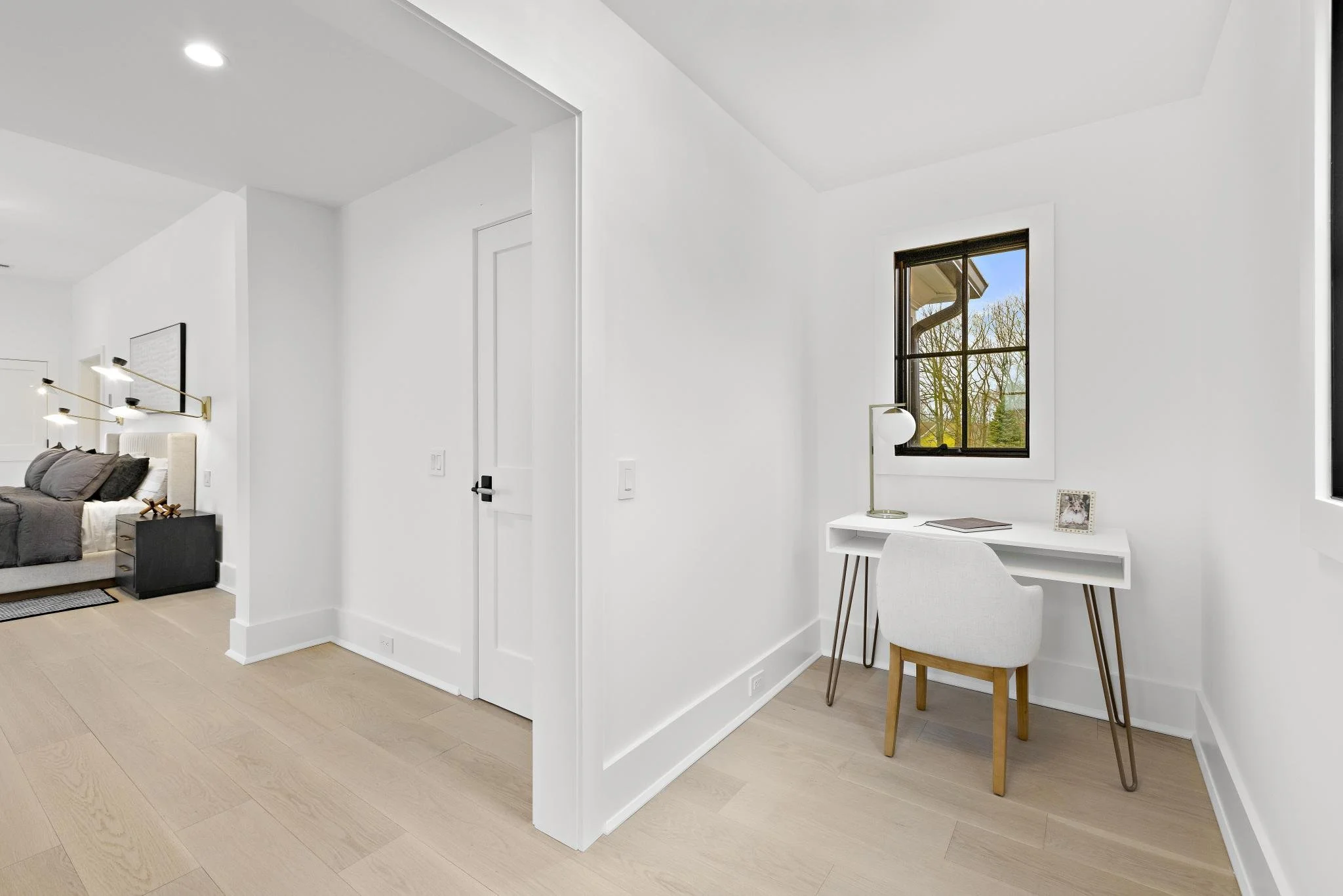 A small white desk with a chair, a lamp, a closed notebook, and a picture frame, situated in a bright corner with a window showing trees outside.