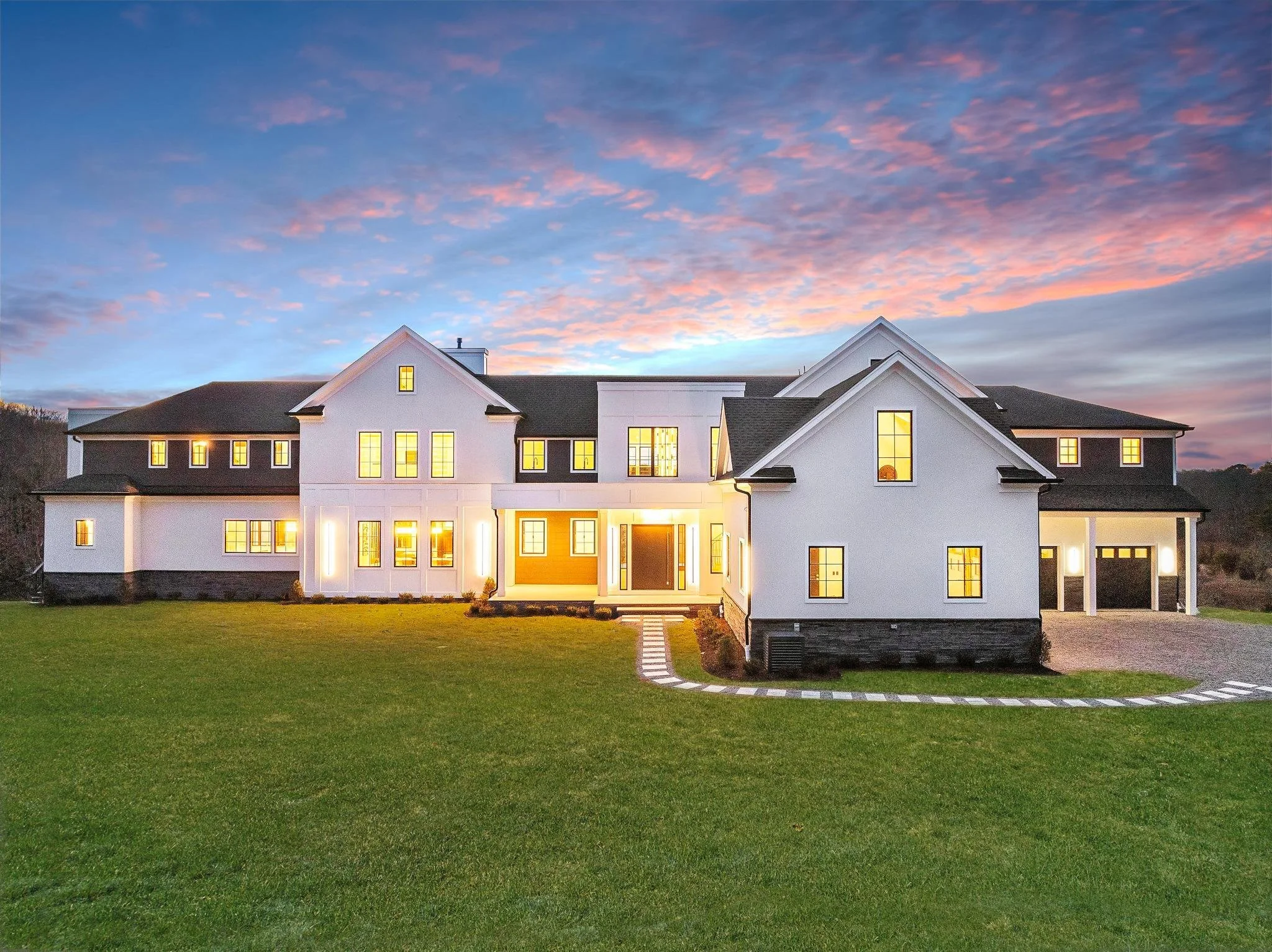 A large, modern white house with multiple gables and a dark roof, illuminated from inside, set against a colorful sunset sky. The house has a well-maintained lawn and a driveway.