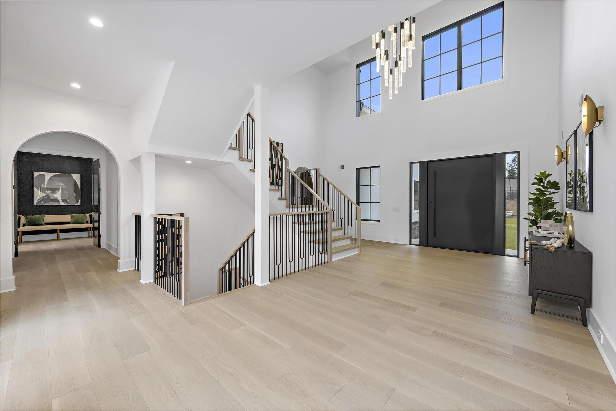 Bright, modern living room with white walls, large black sliding door, light wooden flooring, high ceiling with a chandelier, several windows allowing natural light, and a staircase with black railing leading upstairs. There is a small black console 