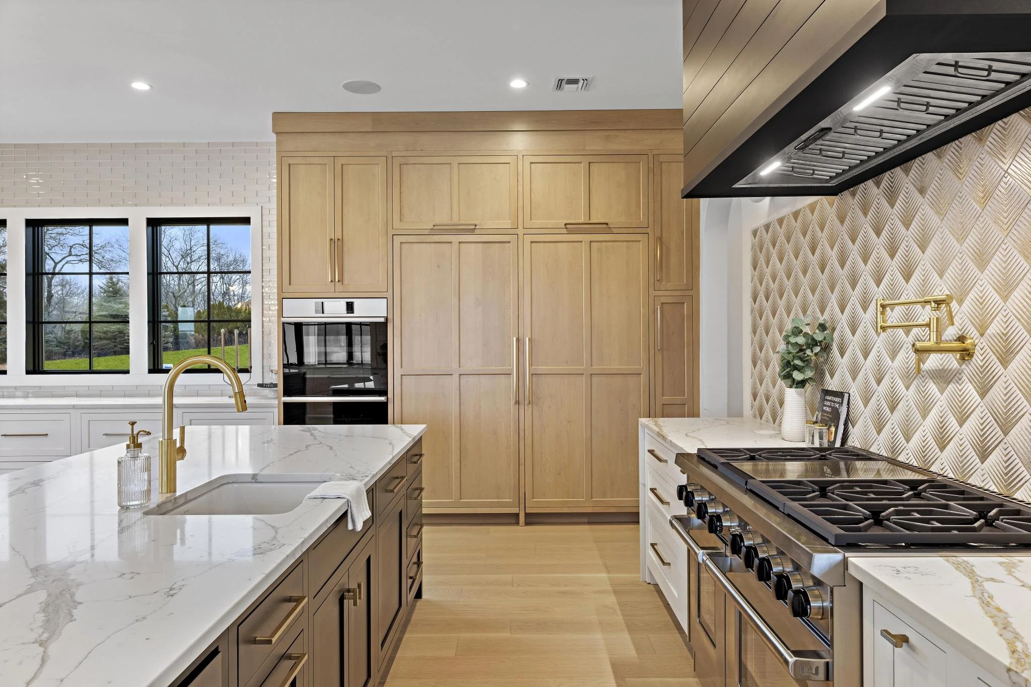 Modern kitchen with white and wood cabinets, marble countertops, a gas stove, gold fixtures, and a window revealing a backyard with trees.