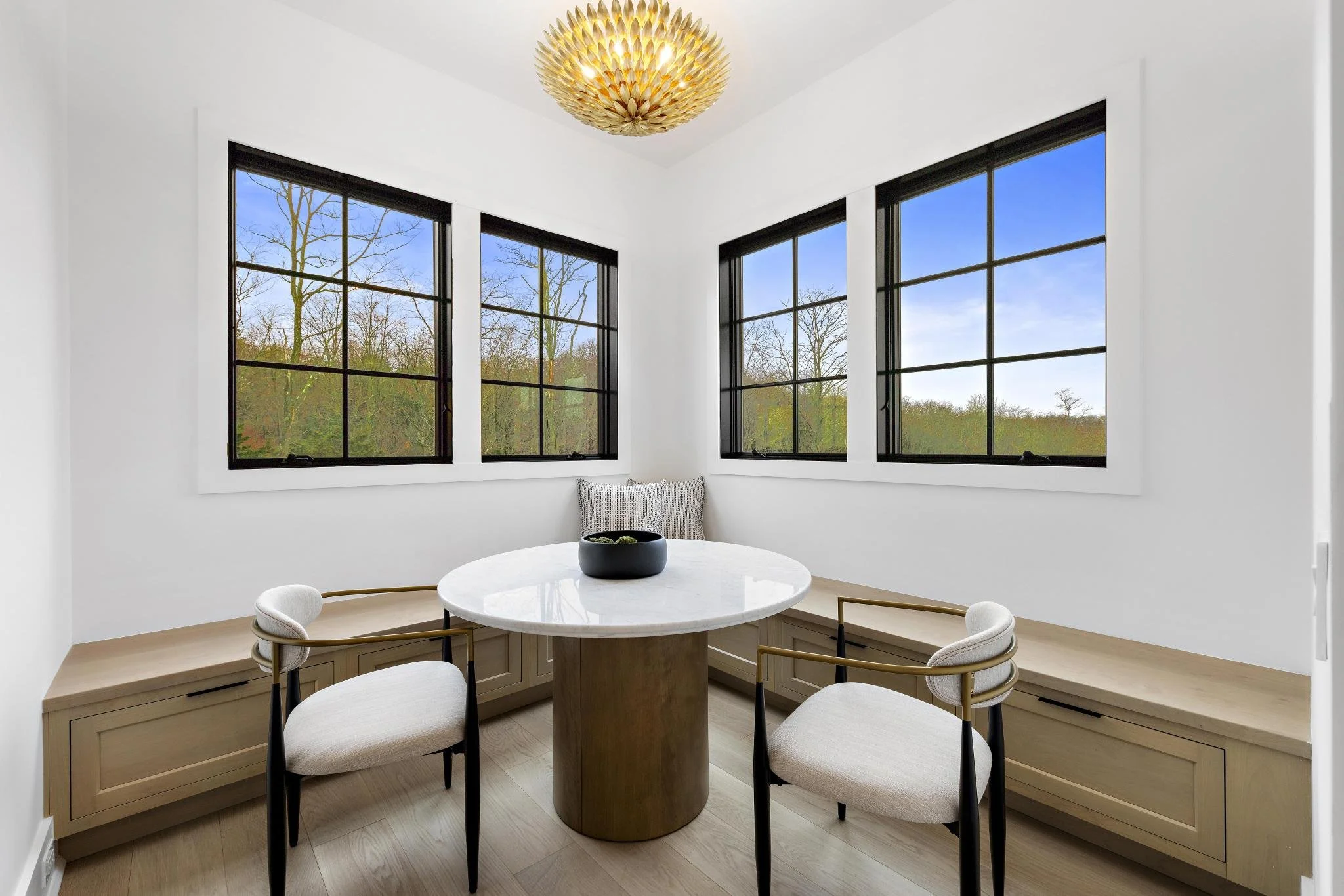Bright dining nook with a round marble table, four modern chairs, built-in wooden bench seating, white walls, and large black-framed windows showing a view of trees outside, with a decorative gold chandelier overhead.