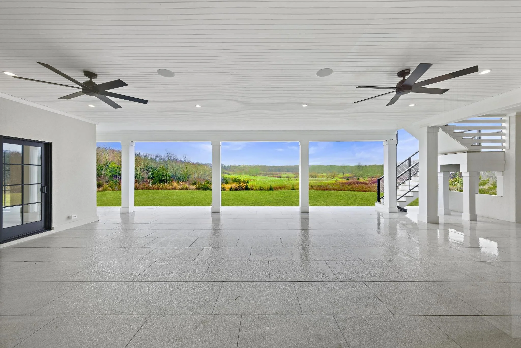 Empty covered porch or patio with white columns, ceiling fans, and large open view of green lawn and distant trees.