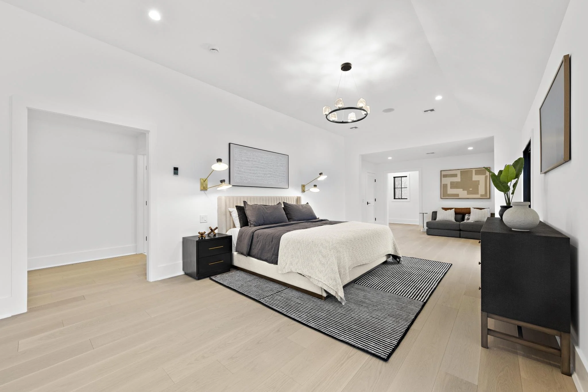 Modern bedroom with king-sized bed, black nightstands, a black rug, grey bedding, a beige upholstered headboard, wall-mounted lamps, a black dresser with vases, and a sitting area with a sofa and artwork on the walls.