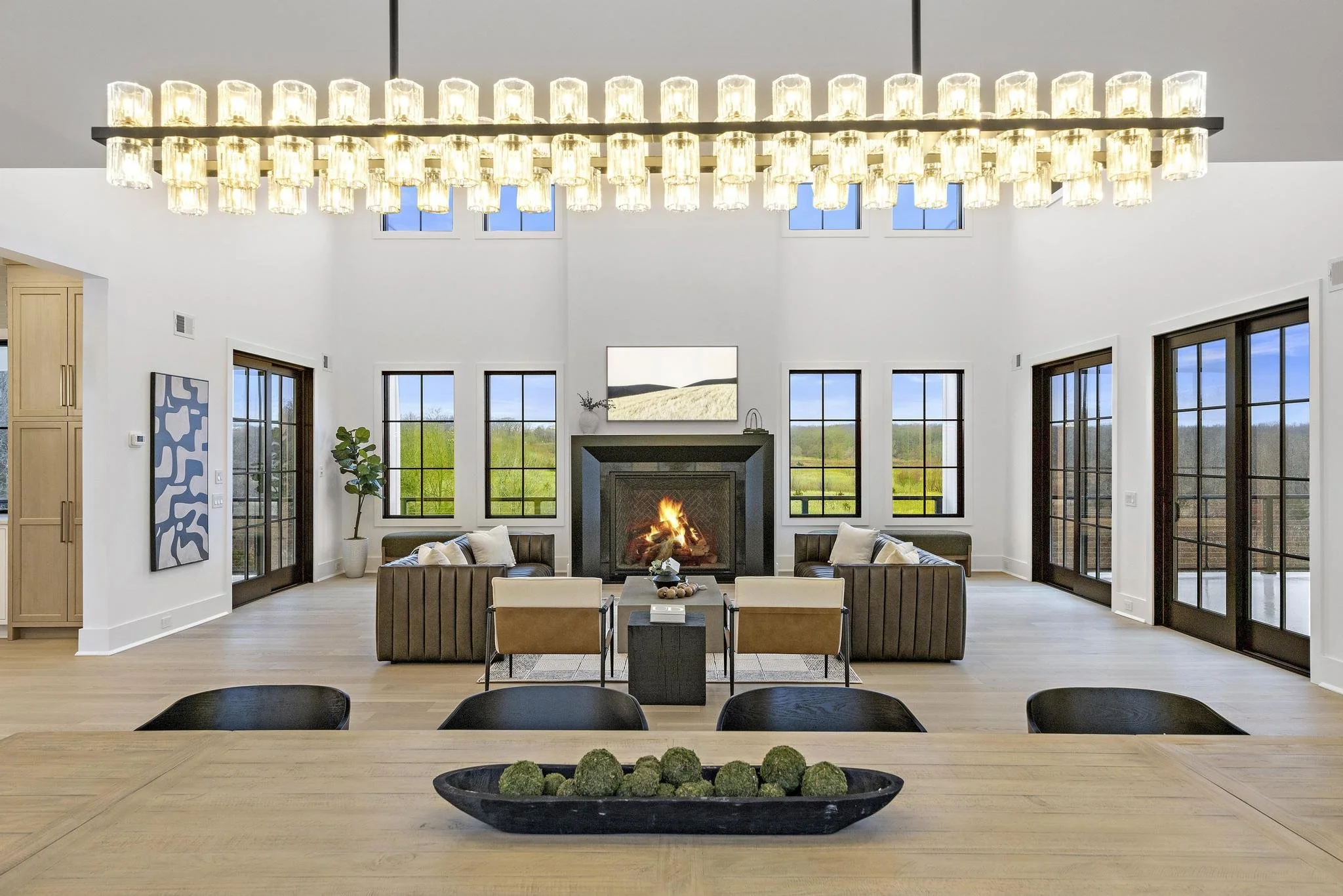Modern living room with black fireplace, six windows showing green landscape, brown sofas, white walls, and a large rectangular chandelier.