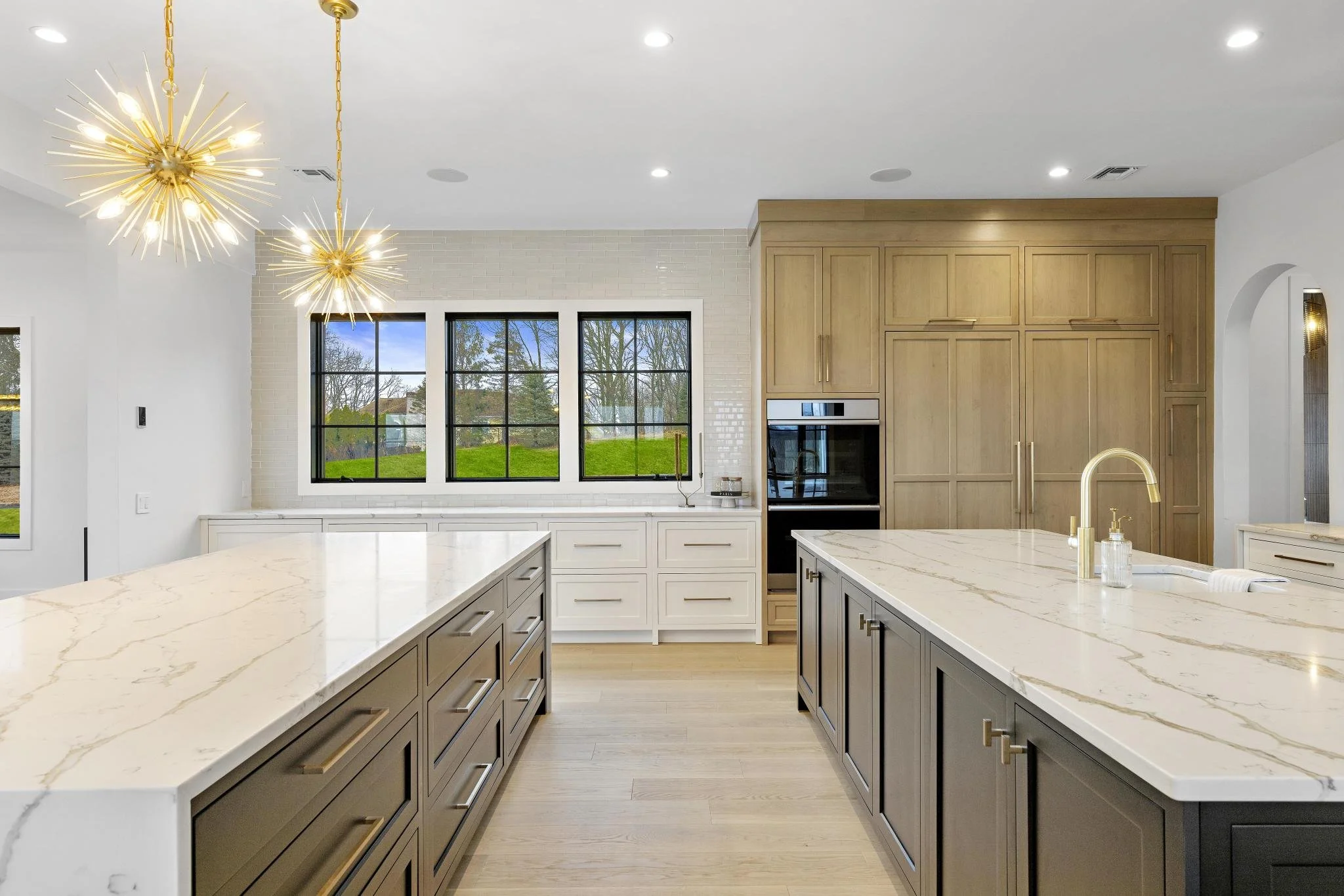 Modern kitchen with two large island countertops with marble surface, dark and white cabinetry, a gold faucet, and three artistic gold chandeliers. Large window showing outdoor greenery.