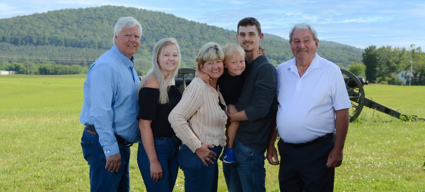 A family of seven standing on a grassy field with mountains in the background, smiling at the camera. They include three older men and women, two younger adults, and two children.