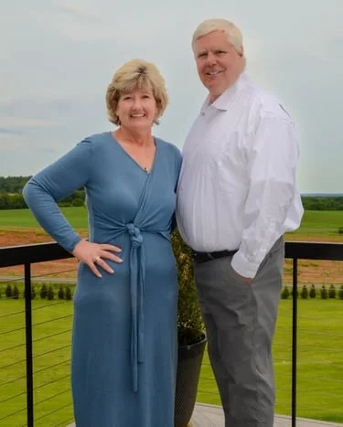 An older woman in a blue dress and an older man in a white shirt standing on a balcony with a scenic green field and cloudy sky in the background, smiling at the camera.
