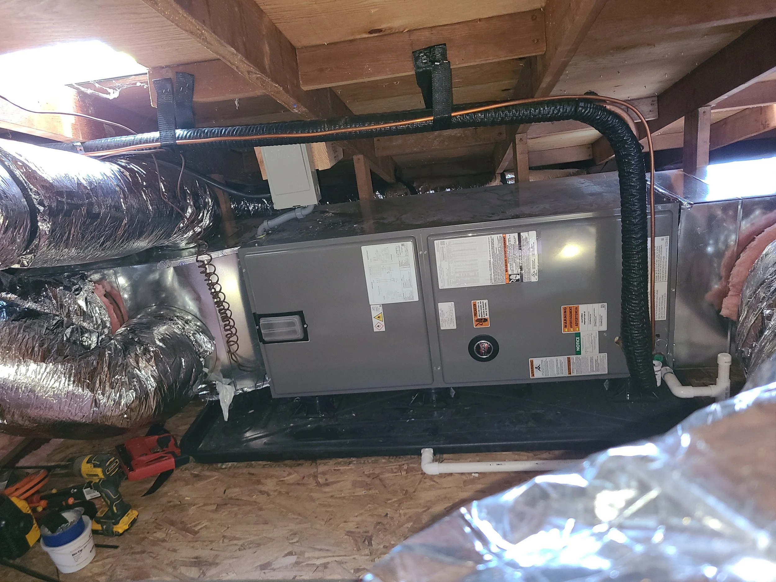 The image shows the interior of an attic with exposed wooden beams and insulation. There is a high-efficiency air handler installed with new ductwork.