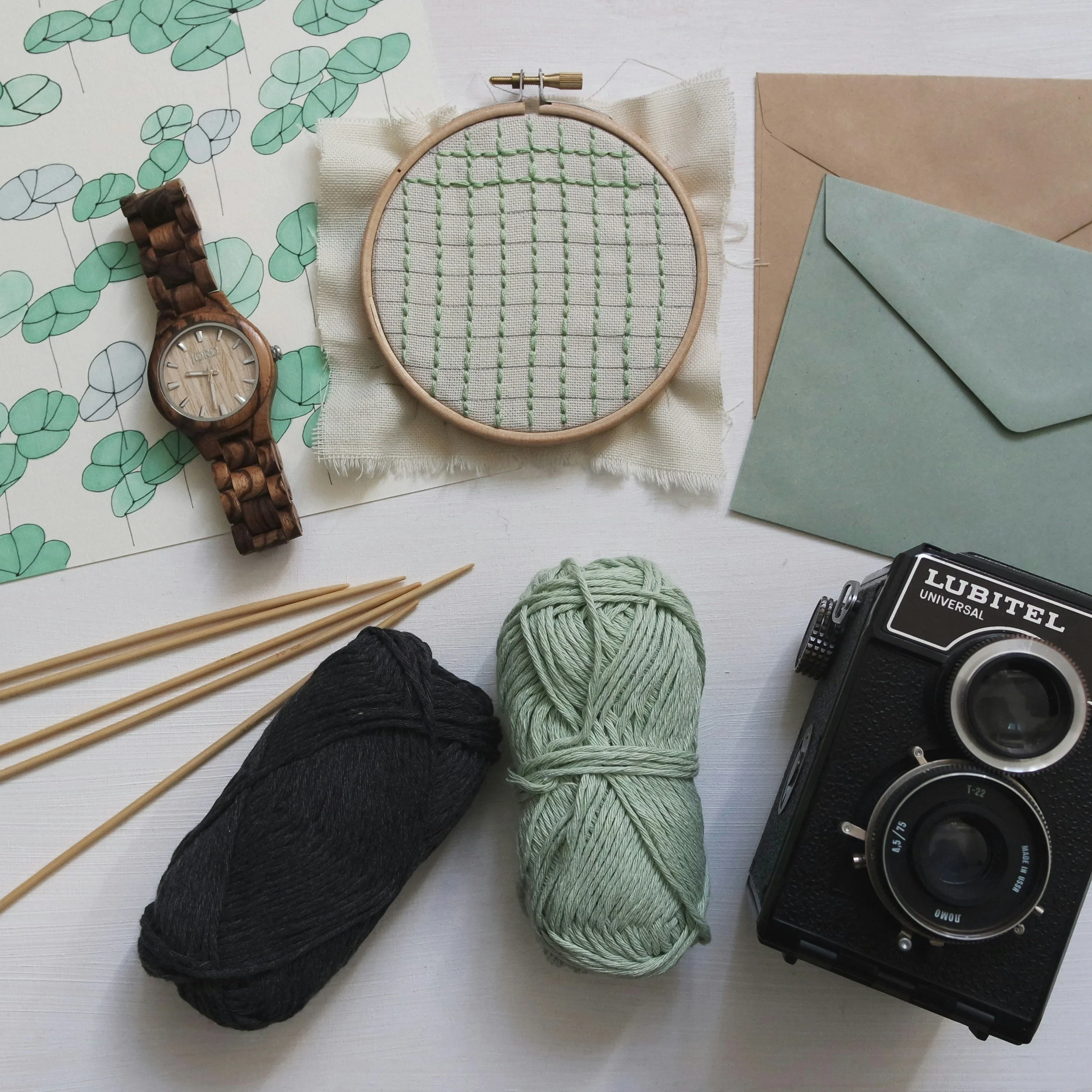 A flat lay of crafting and stationery items including a wooden watch, an embroidery hoop with fabric and green stitches, envelopes, yarn in black and light green, knitting needles, a black vintage camera, and a floral print paper.