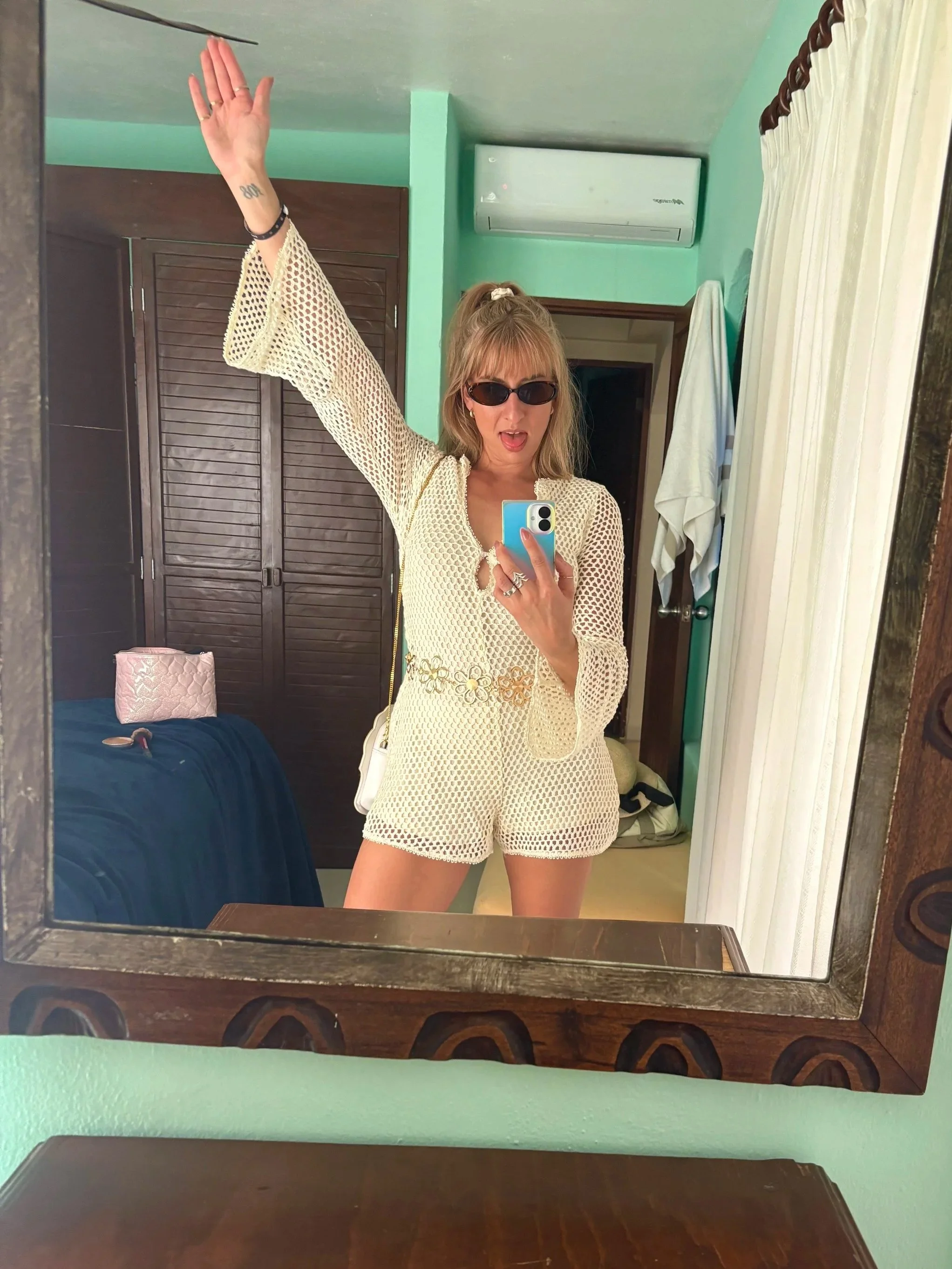 A woman in sunglasses taking a mirror selfie in a room with green walls, wearing a cream-colored crochet outfit, with her left arm raised, and blonde hair tied back.