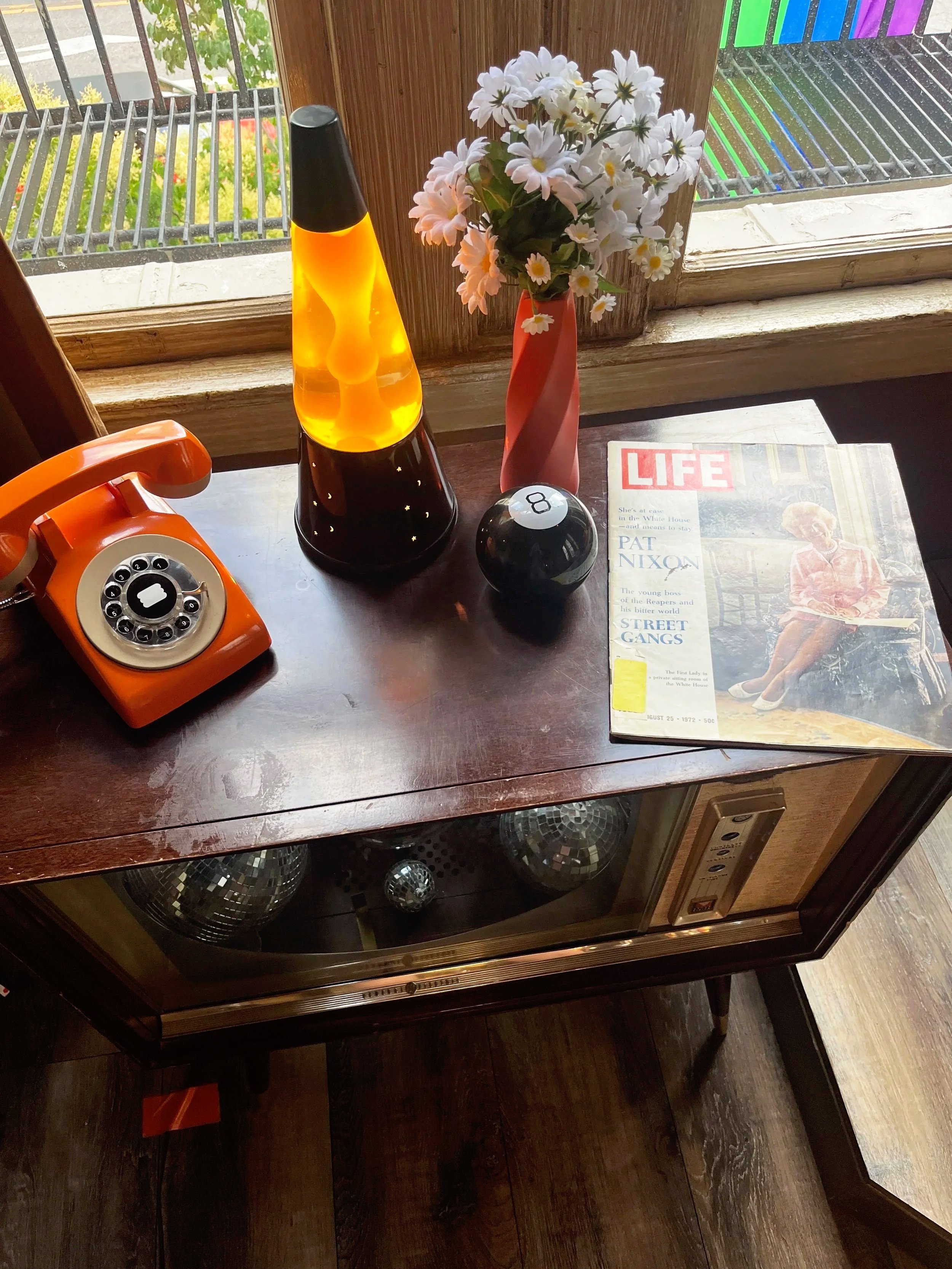 A vintage orange rotary phone, a lava lamp, a black 8-ball, a vase with white daisies, a Life magazine featuring Pat Nixon on the cover, and a window with a view of a garden and a fence.