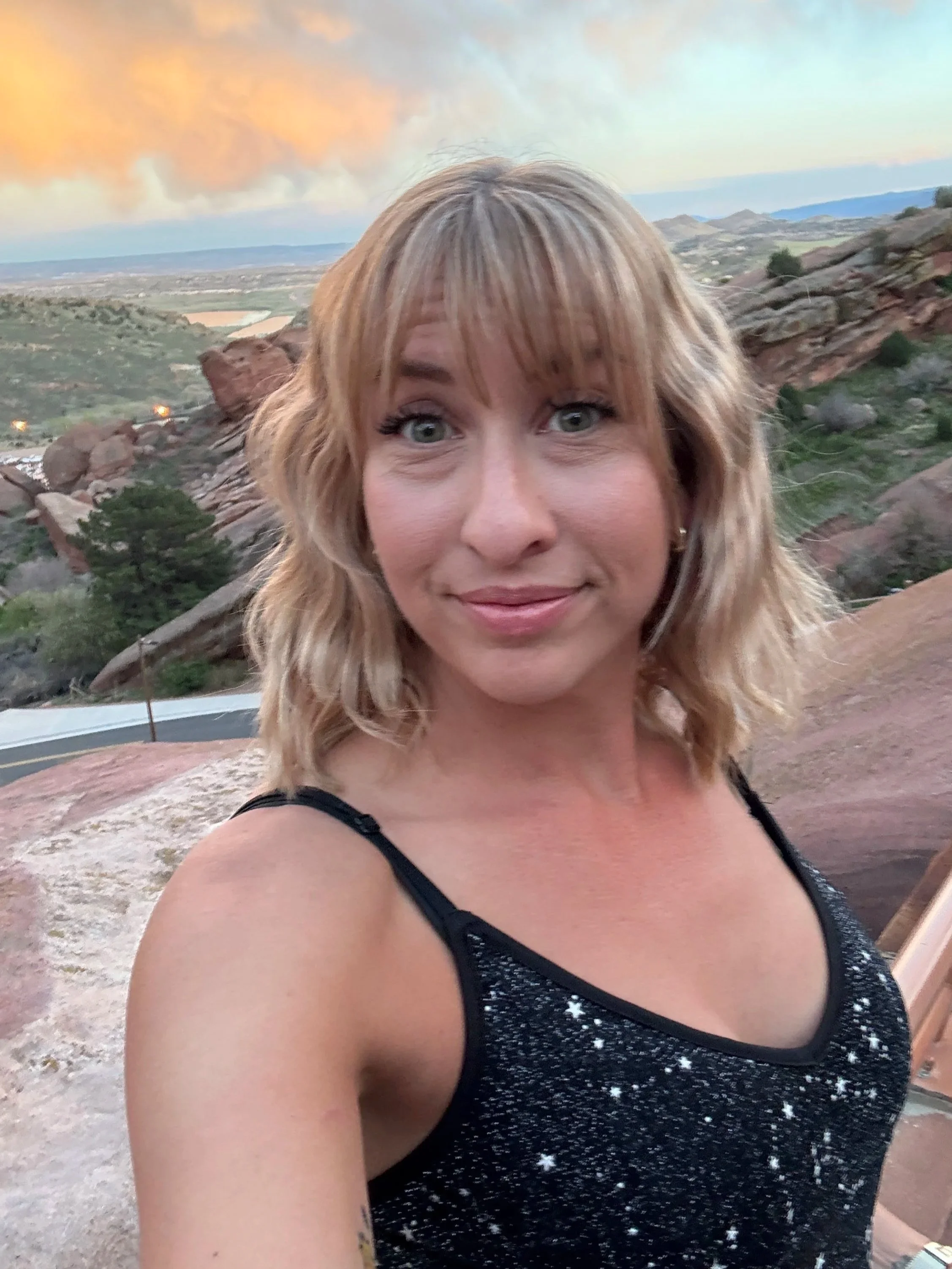 A woman taking a selfie outdoors with a rocky landscape and sunset sky in the background.