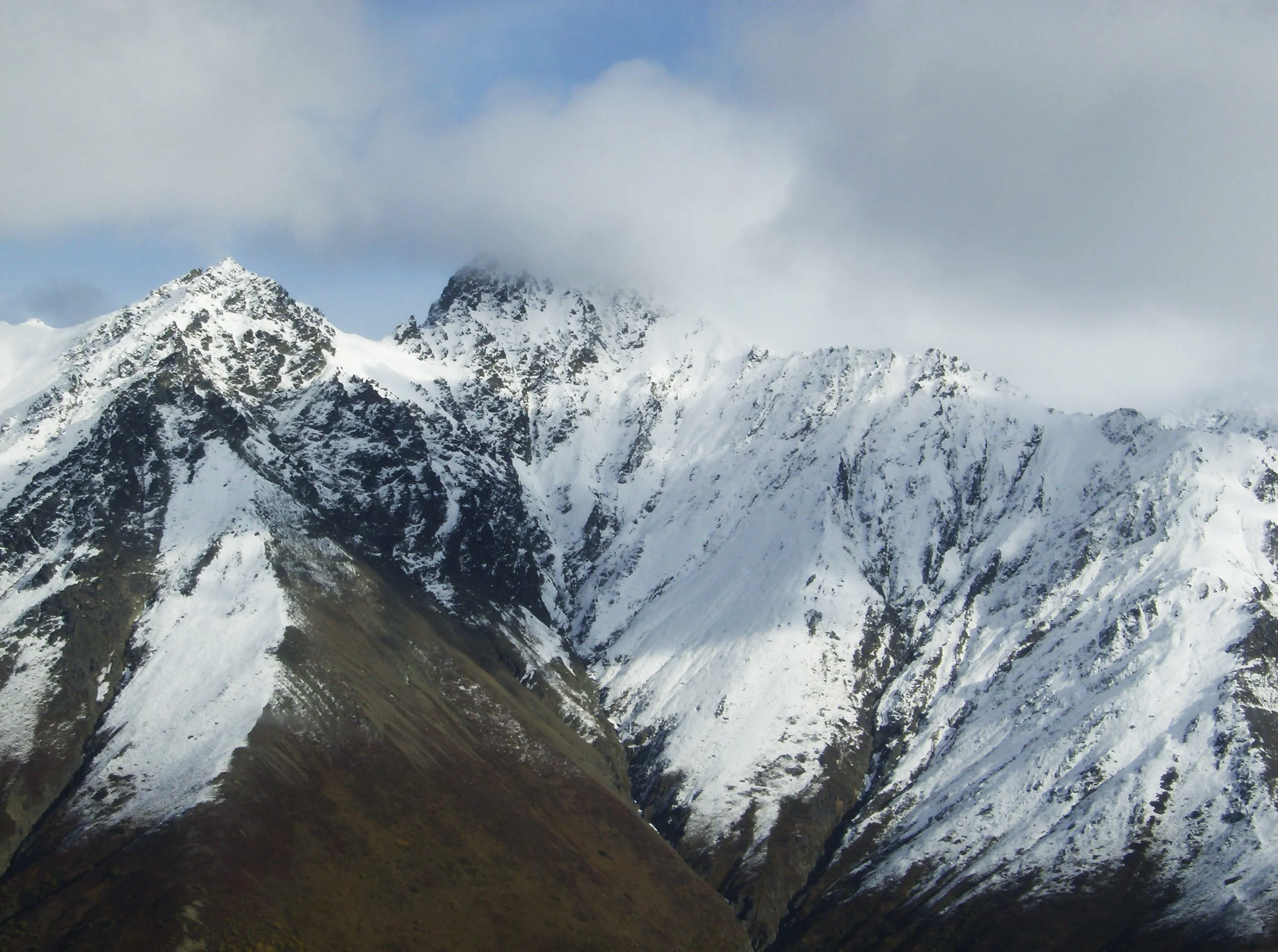 Snow-covered mountain peaks partially covered by clouds under a blue sky.