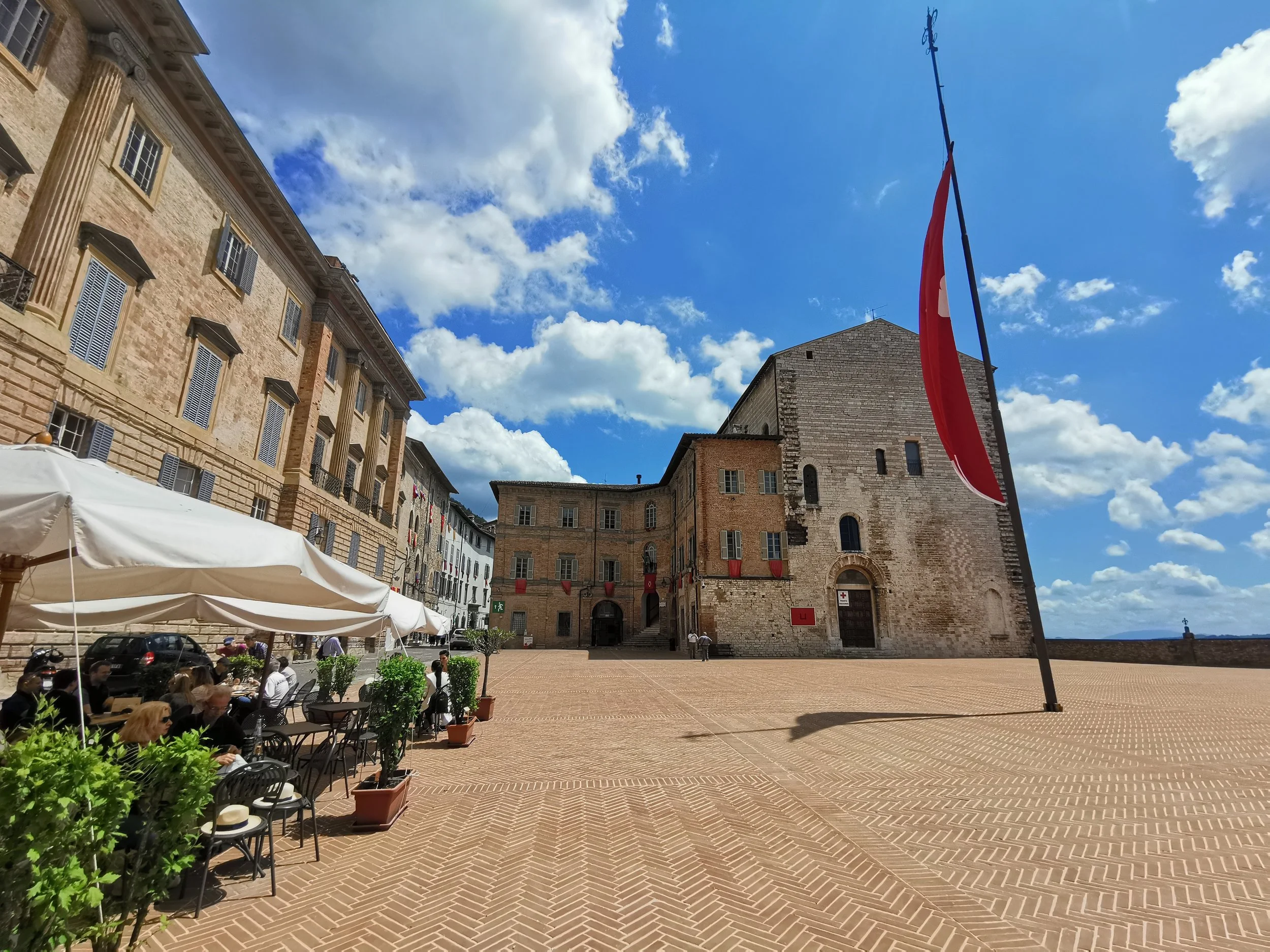 A historic stone building in an open square in Gubbio, Italy with outdoor seating and umbrellas, under a blue sky with scattered clouds.