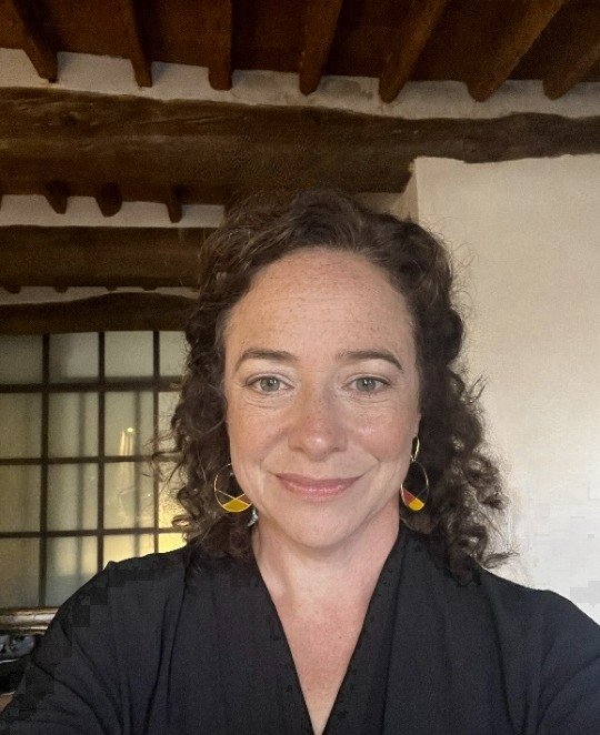 A woman with curly brown hair wearing earrings and a black top, smiling, indoors with wooden beams on the ceiling and a grid window in the background.