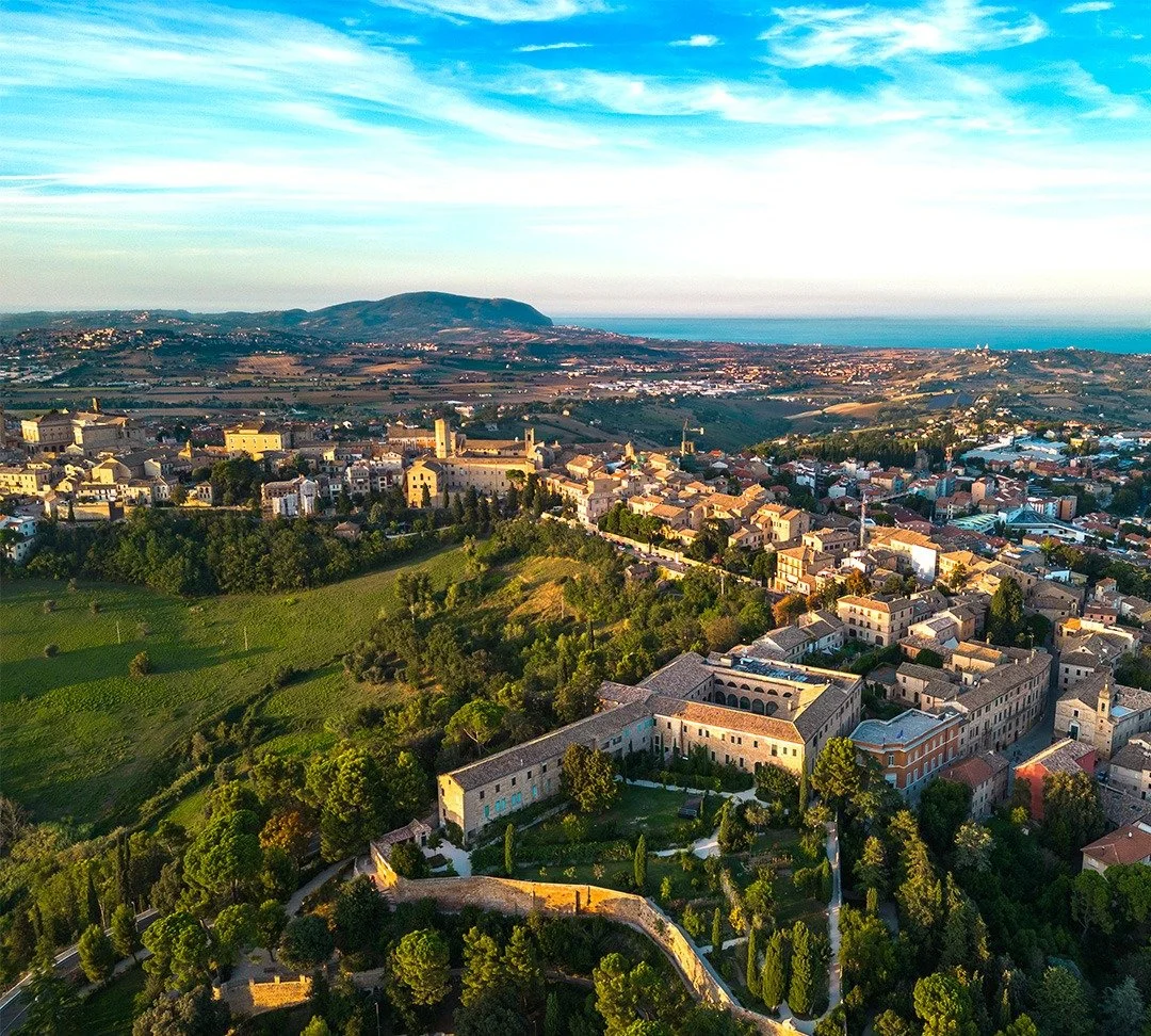 Aerial view of Recanati, Italy with green hills, historic buildings, and the ocean in the background under a blue sky with scattered clouds.