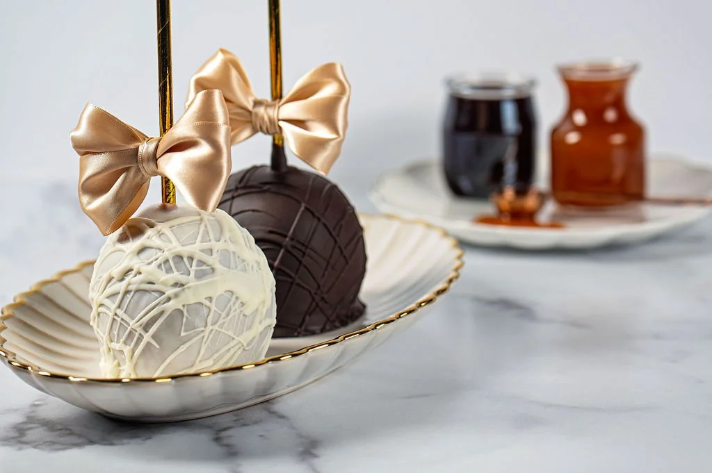 Three decorated cake pops with gold bows in a white and gold dish, with jars of chocolate and caramel in the background.