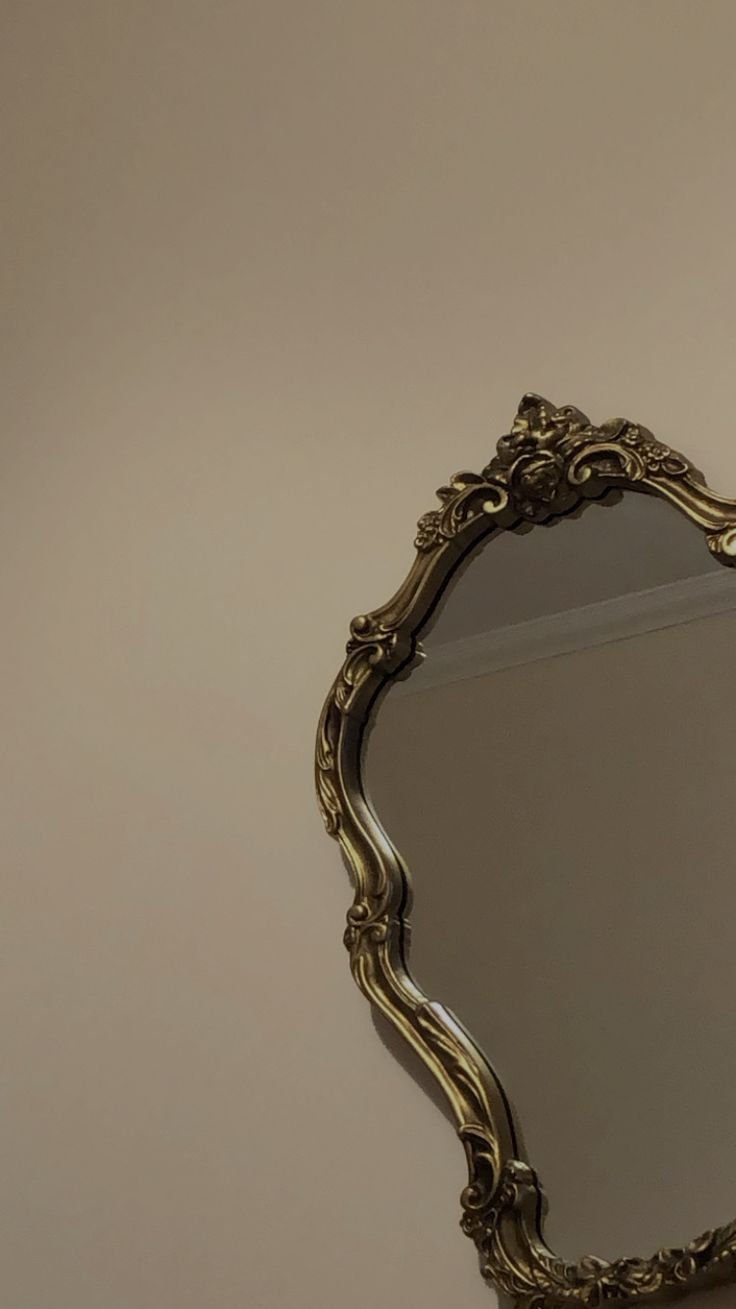 Image of ornate, gold-colored mirror with elaborate carved frame hanging on a beige wall.