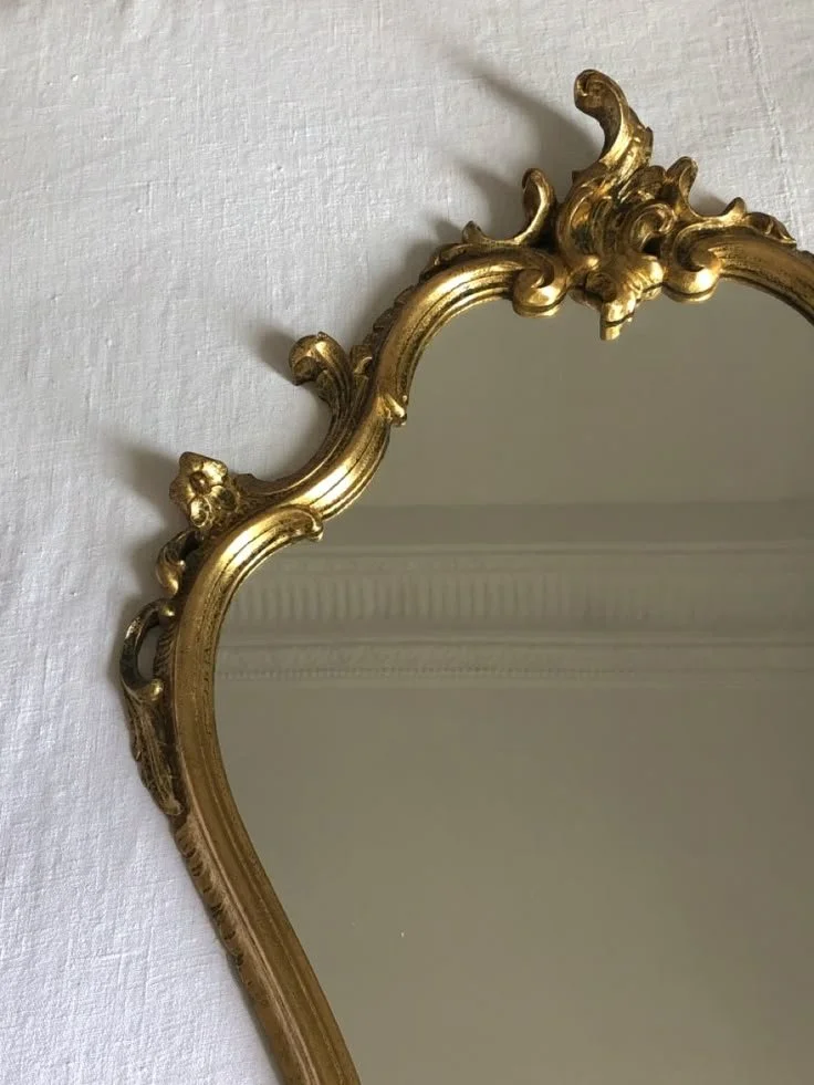 Close-up of an ornate, gold-framed mirror hanging on a white wall.